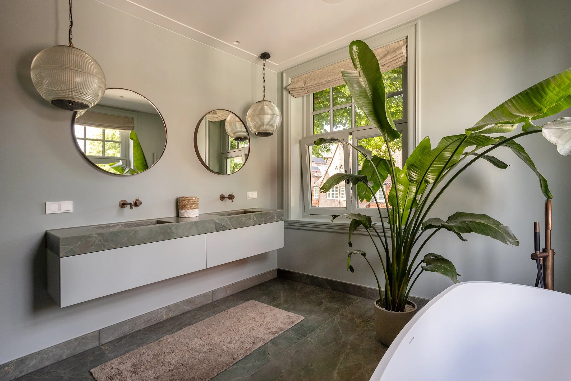 Een moderne badkamer met twee ronde spiegels, hangende lampen, een groene natuurstenen wastafel, een grote plant in een pot, en een raam met een uitzicht op een tuin. Decoratie en natuurlijke lichtinval accentueren de ruimte.