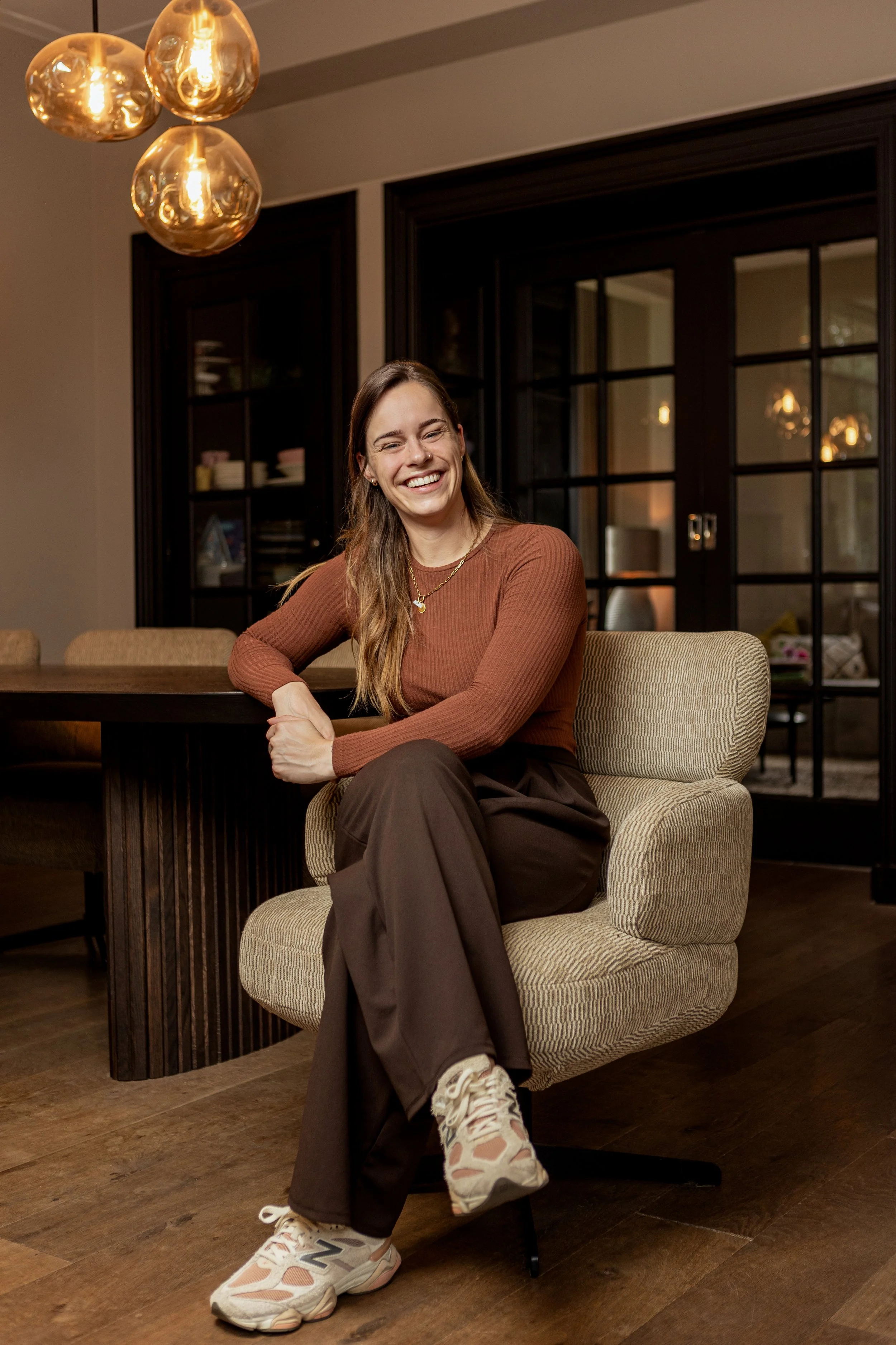Een lachende vrouw zit op een beige gestoffeerde stoel met een houten vloer op de achtergrond, in een gezellig interieur met zwarte glazen deur en gouden hanglampen.