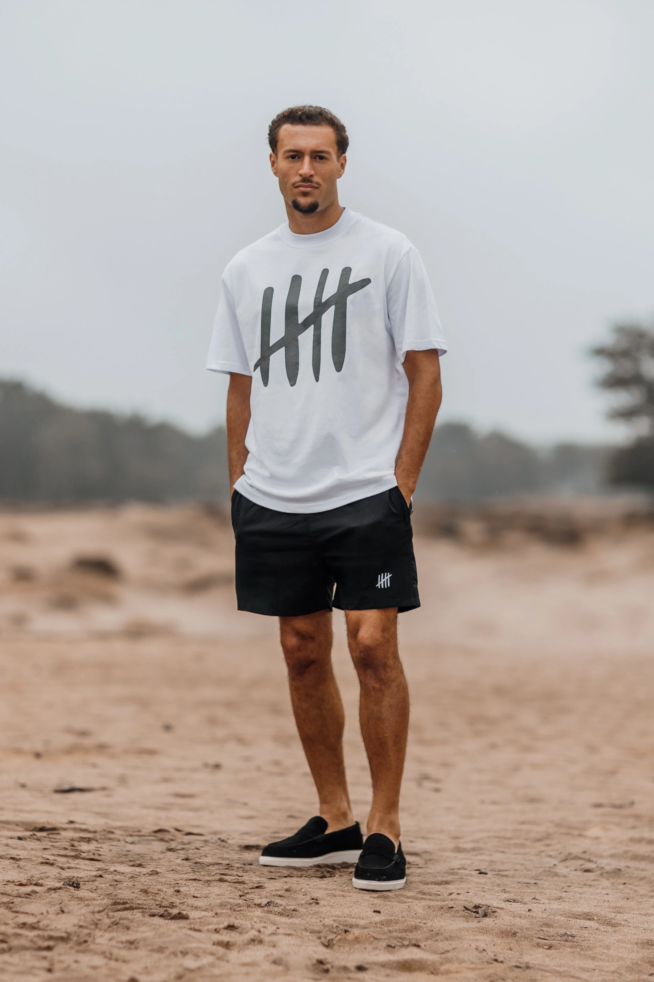 Man staat op een zandstrand, draagt een wit T-shirt met een talloosheidsprint en korte zwarte broek, en kijkt naar de camera.