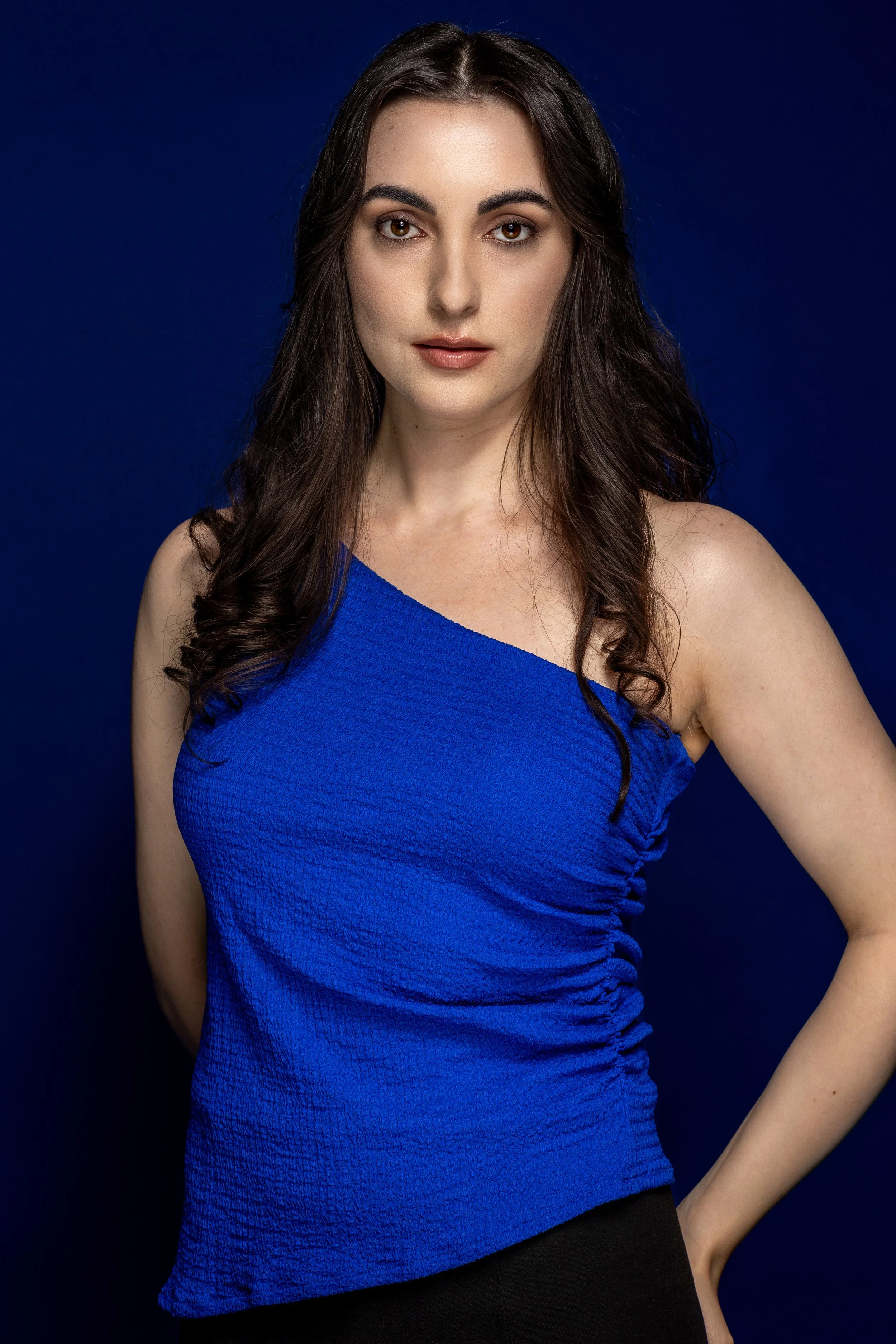 Portret van een jonge vrouw met lang donker haar, witte huid en bruin ogen, draagt een blauwe een-schouder top en poseert voor een donkere blauwe achtergrond.