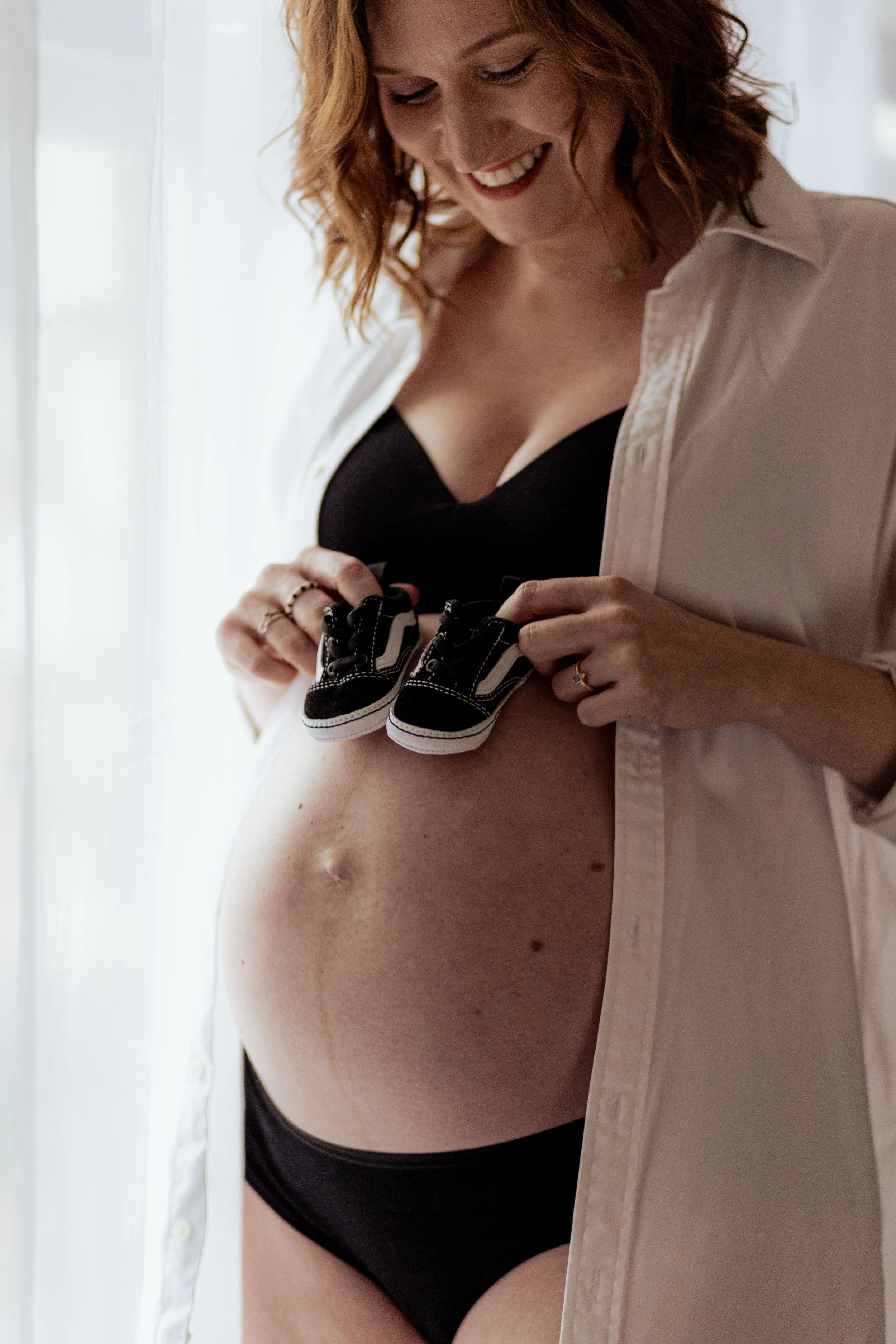 Zwangere vrouw met rood haar houdt kleine zwarte sneakers tegen haar buik, glimlachend.