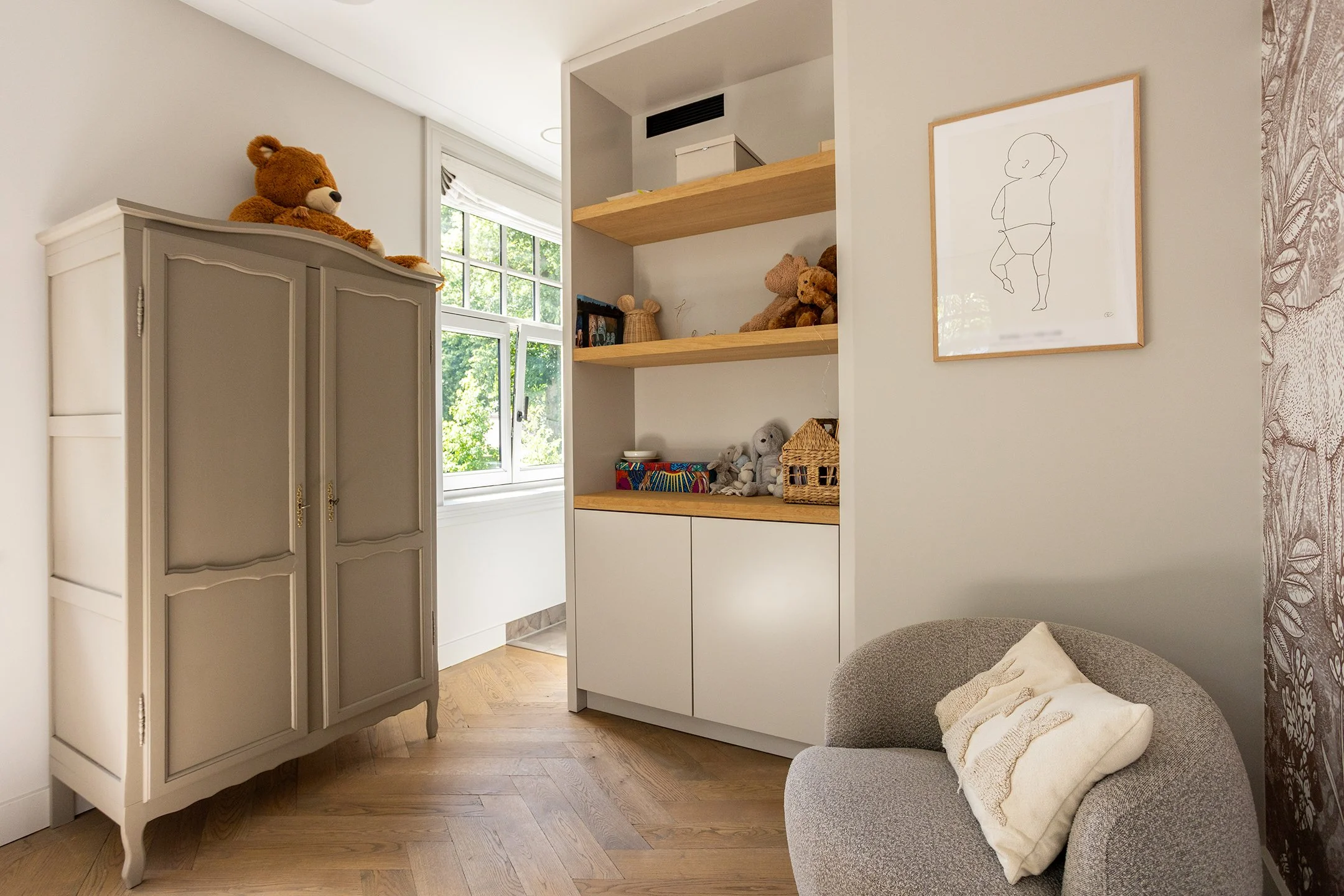 Interieur van een kinderkamer met een beige kast met een teddybeer, een witte ladekast met teddyberen en speelgoed, een raam met uitzicht op groene bomen, een paar witte pluchen knuffels op een plank, een grijze stoel met een beige kussen, en een muu
