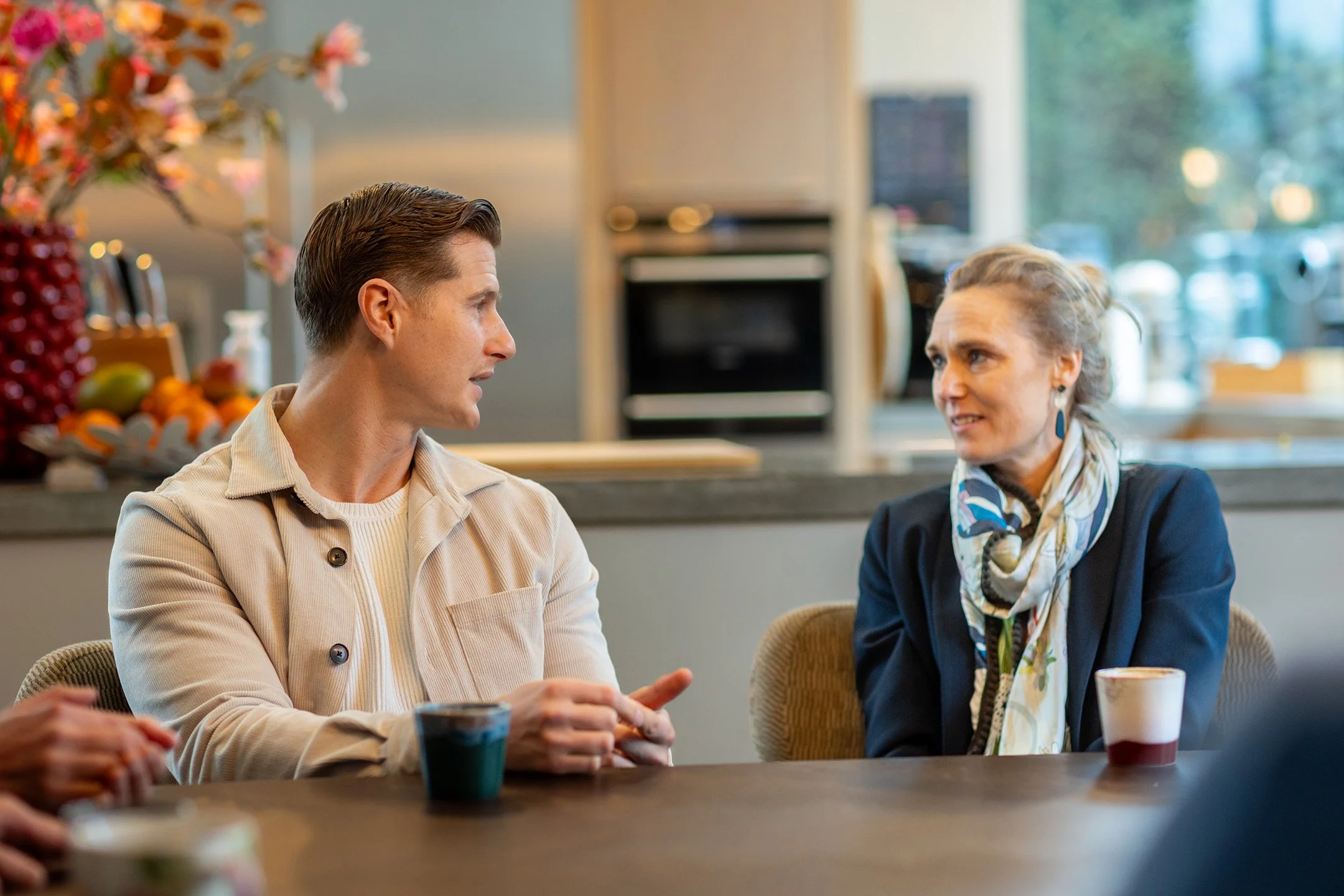 Twee mensen die in een keuken met een ontbijtbar aan een tafel zitten, betrokken in een gesprek.