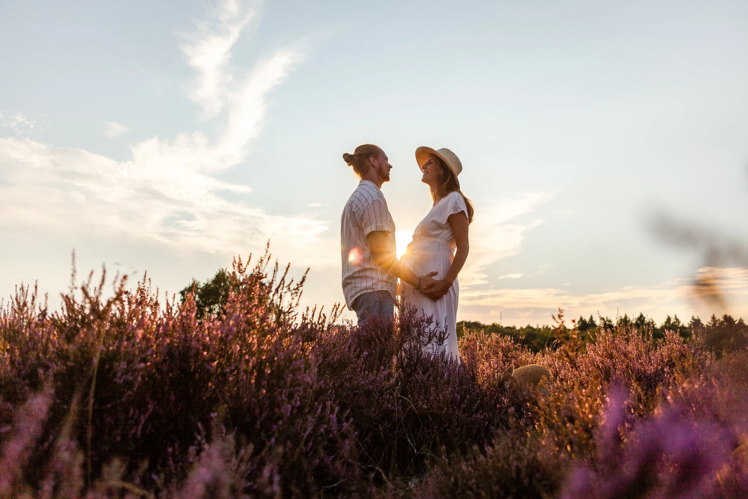 Een zwangere vrouw en haar partner staan in een veld met bloemen tijdens zonsondergang, houden elkaars handen vast en kijken elkaar lieflick.