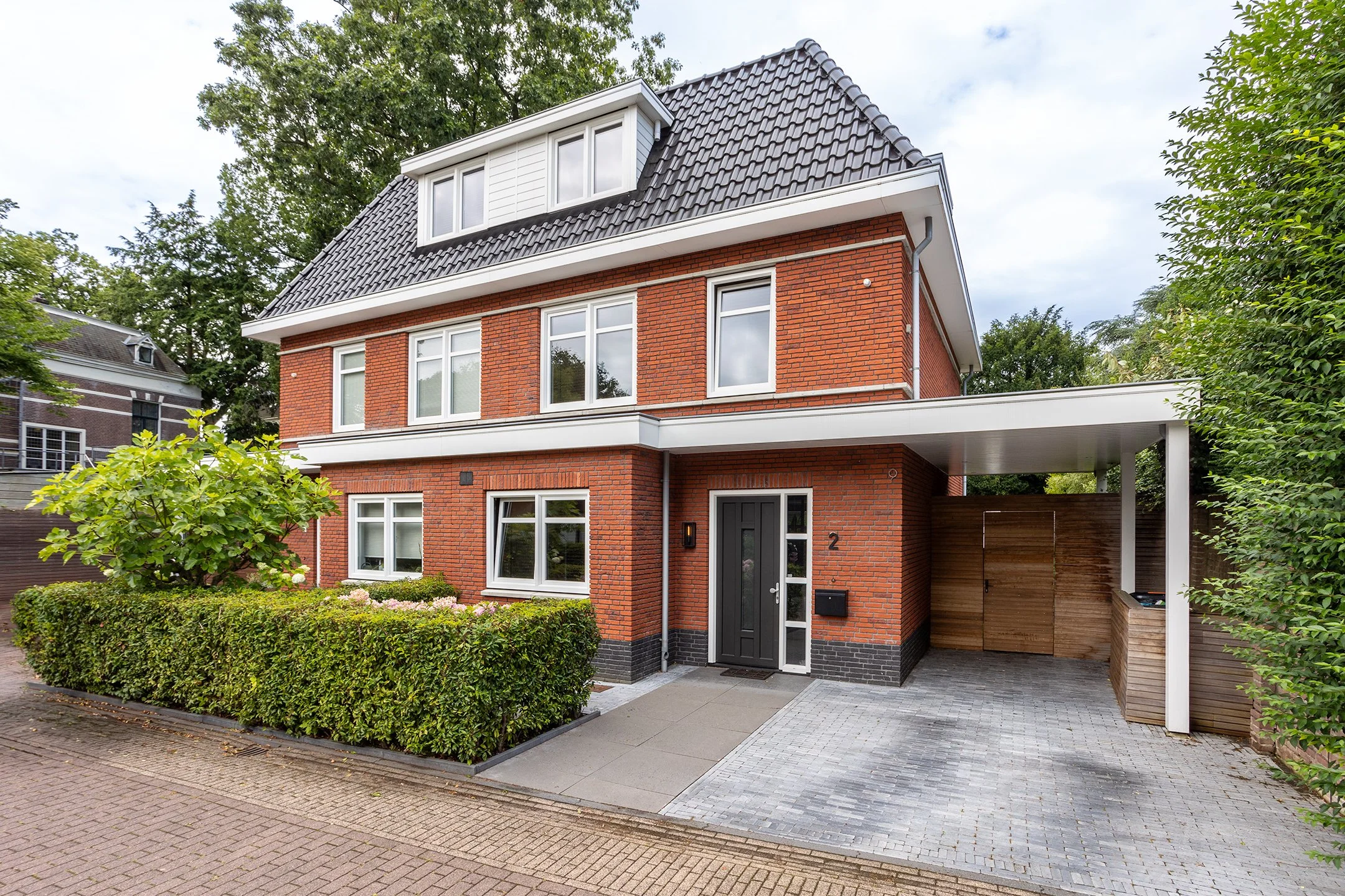 Een modern, vierlaagse woning gemaakt van rood baksteen met een zwarte voordeur en grote ramen. Het huis heeft een overkapping en een oprit met stenen tegels.