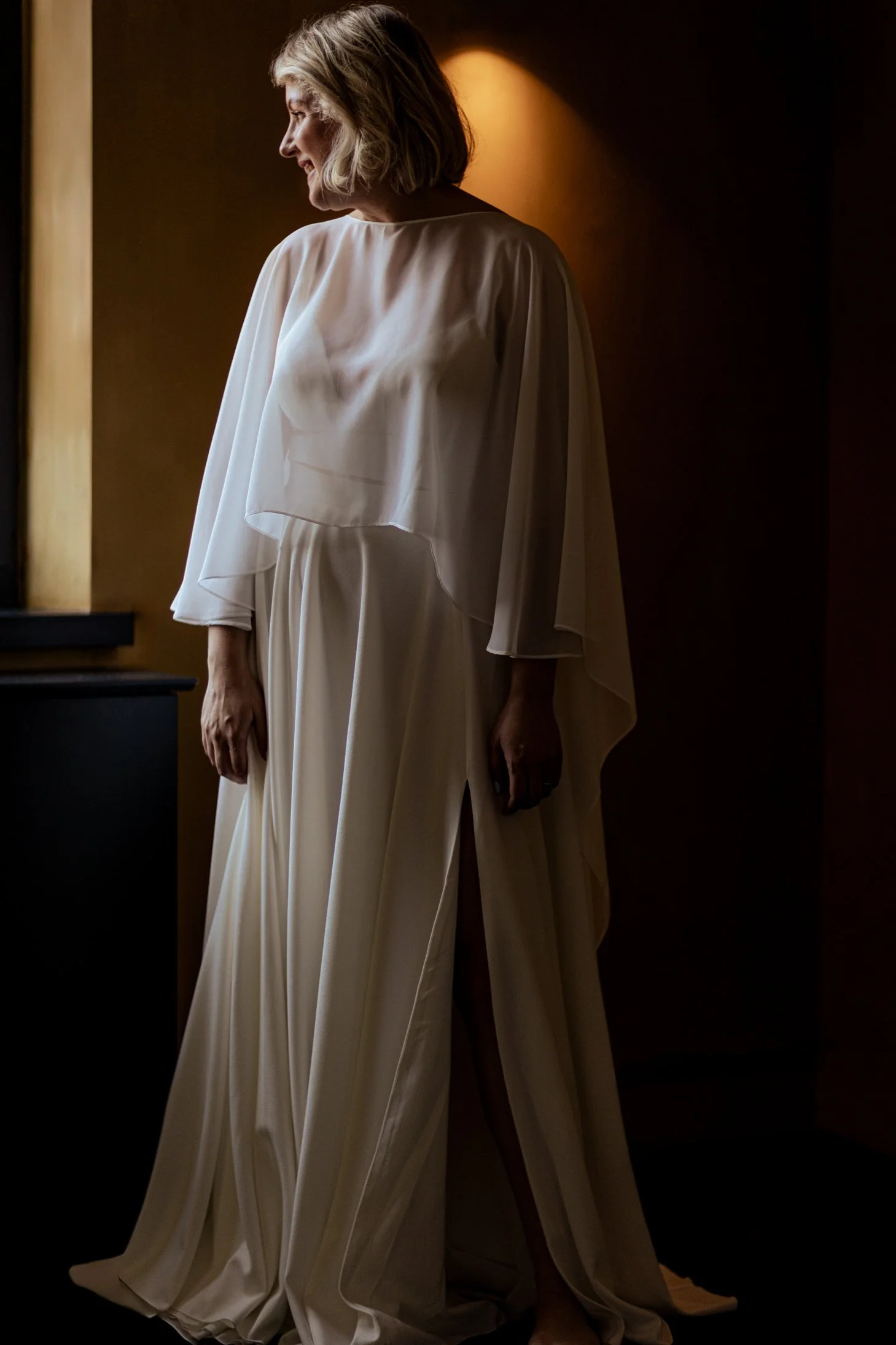 Een oudere vrouw in een witte, vloerlange jurk met wijde mouwen staat in een dim verlichte kamer, haar hoofd is naar links gericht.