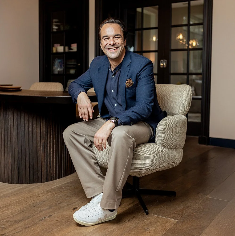 Een man die lachend op een beige bureaustoel zit in een kantoor, gekleed in een blauw jasje en beige broeken, met witte sneakers.