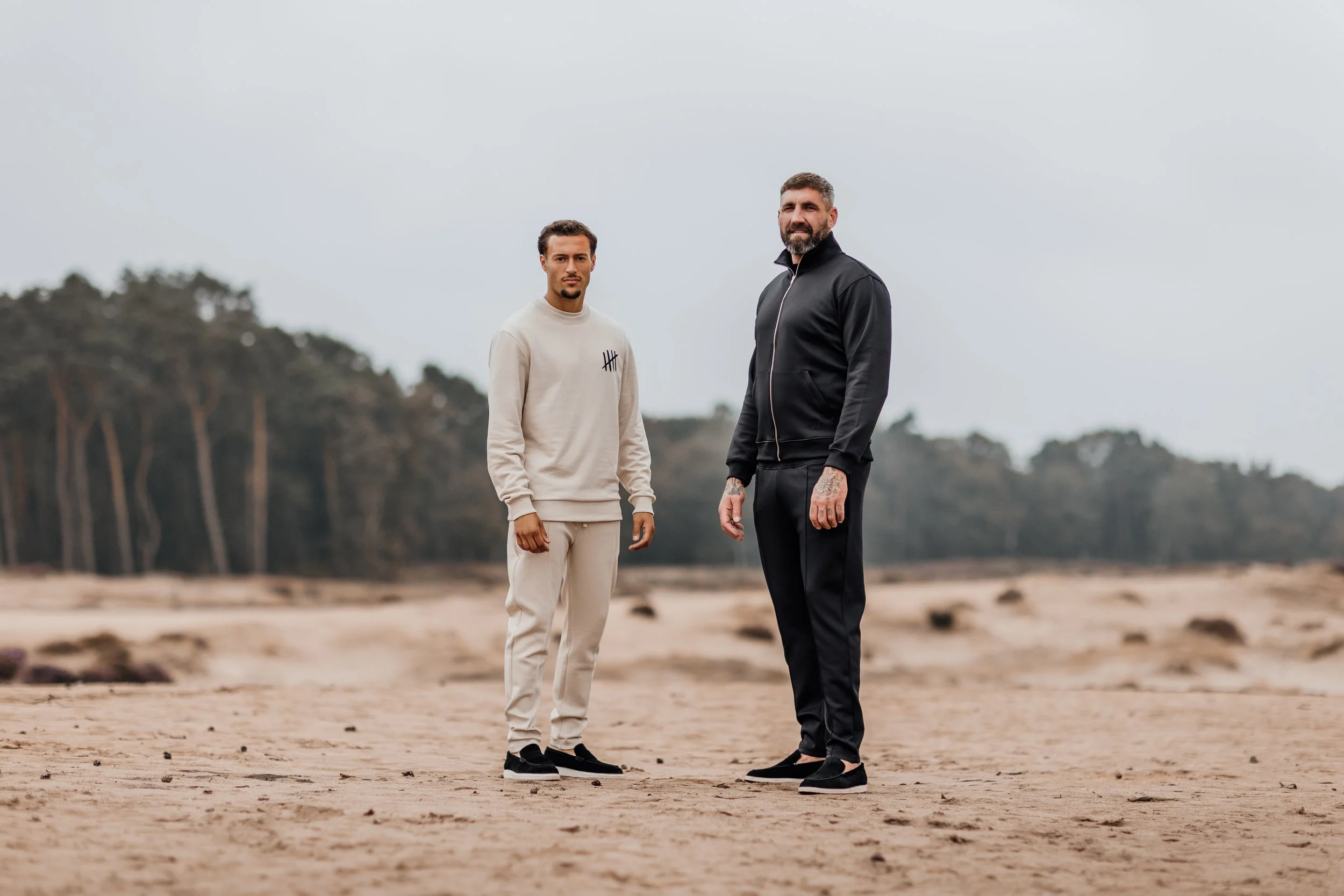 Twee jongens staan op een zandstrand, met een bos in de achtergrond, gekleed in casual sportkleding.