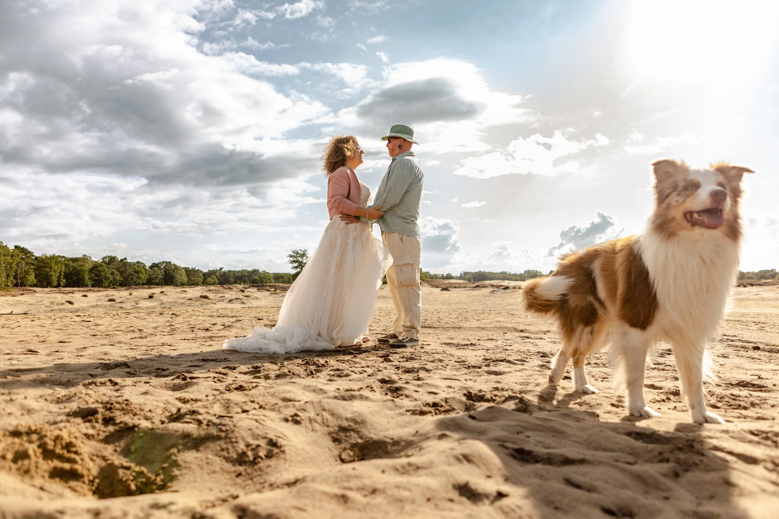 Een bruid en bruidegom staan op een zandstrand onder een bewolkte hemel, ze kijken elkaar liefdevol aan, met een hondenras die vrolijk op de voorgrond staat.