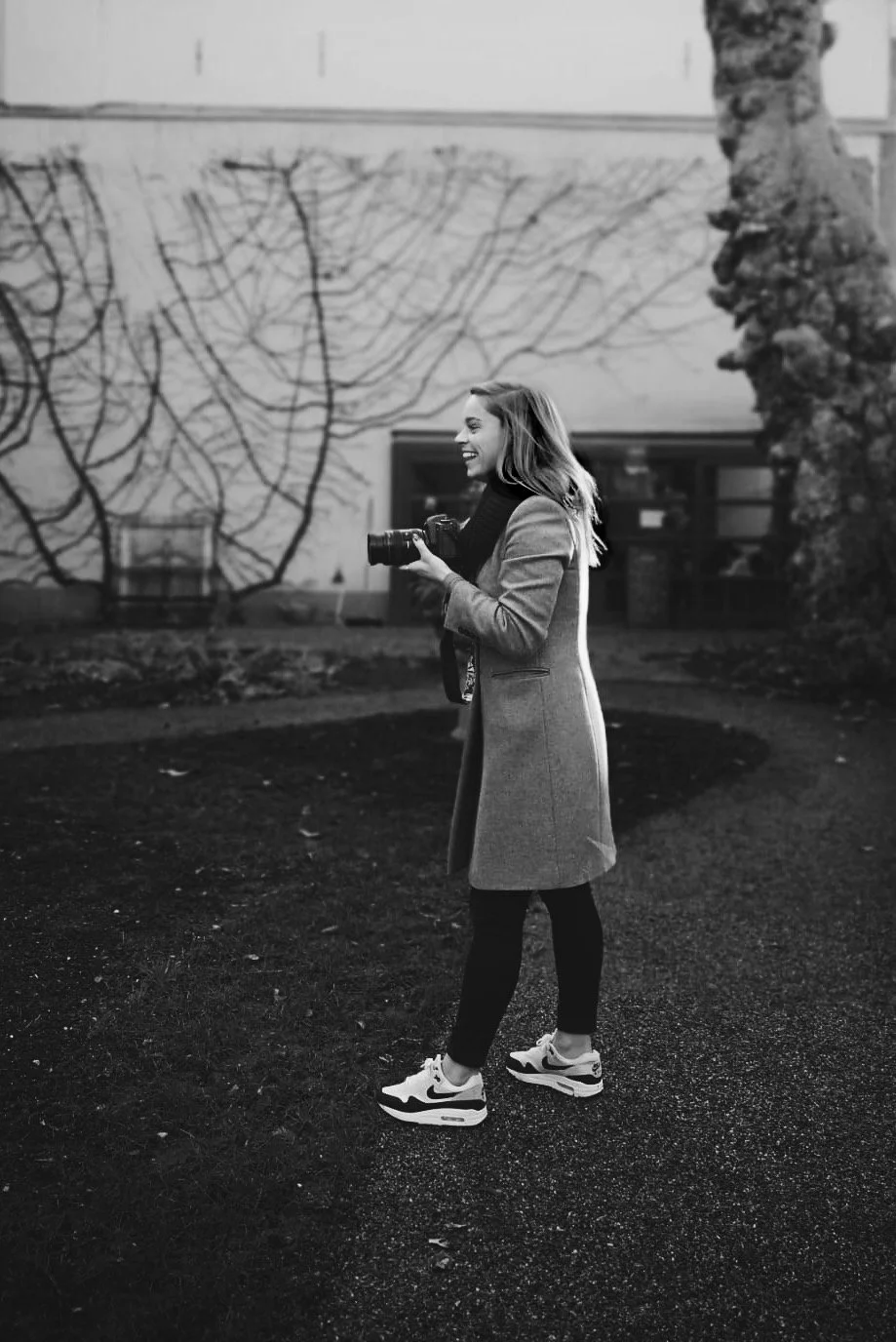 Een lachende vrouw in een jas met een camera in haar handen, grote sneakers, buiten op een grasveld met een muur en boom in de achtergrond.