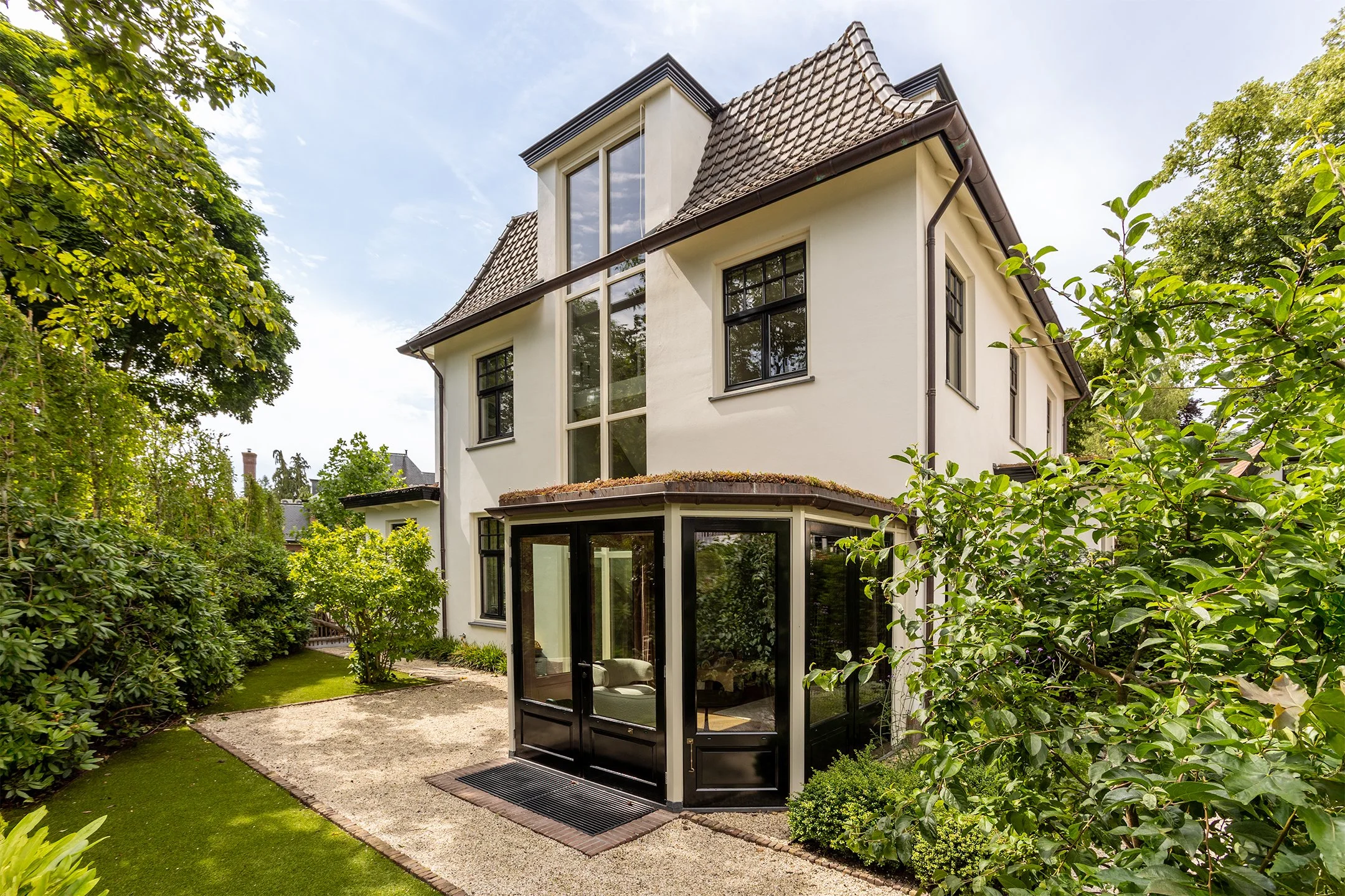 Een wit huis met meerdere verdiepingen en grote ramen, omgeven door groene bomen en een tuin met een zandpad en gras.