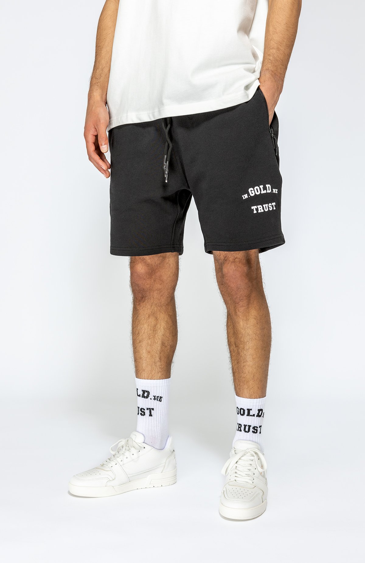 Man met witte sneakers, witte sokken met 'GOLD' en 'TRUST', zwarte shorts met 'GOLD' en 'TRUST', en een witte T-shirt.