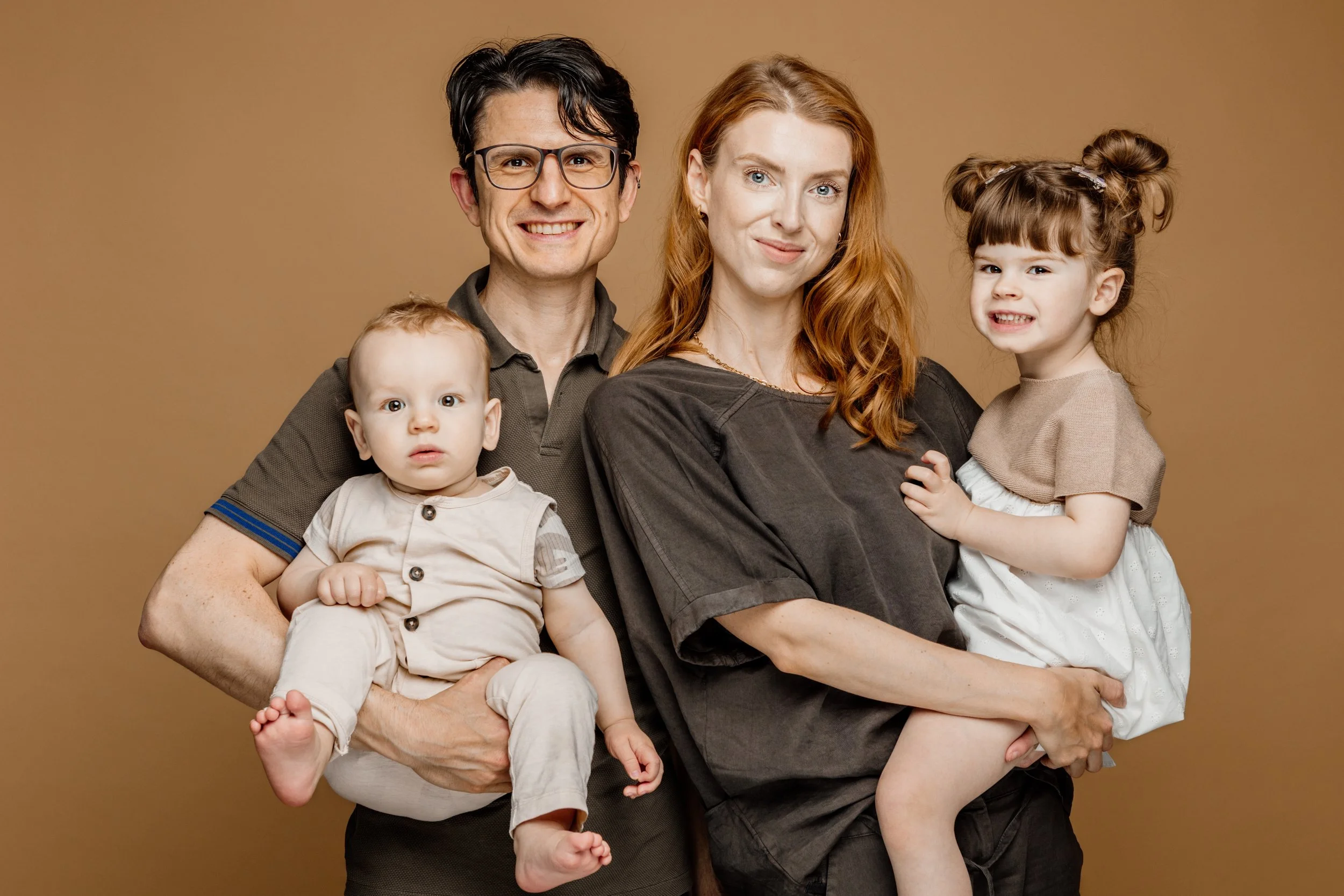 Gezicht van een gezin met vader, moeder, een baby en een peuter, staand voor een beige achtergrond.