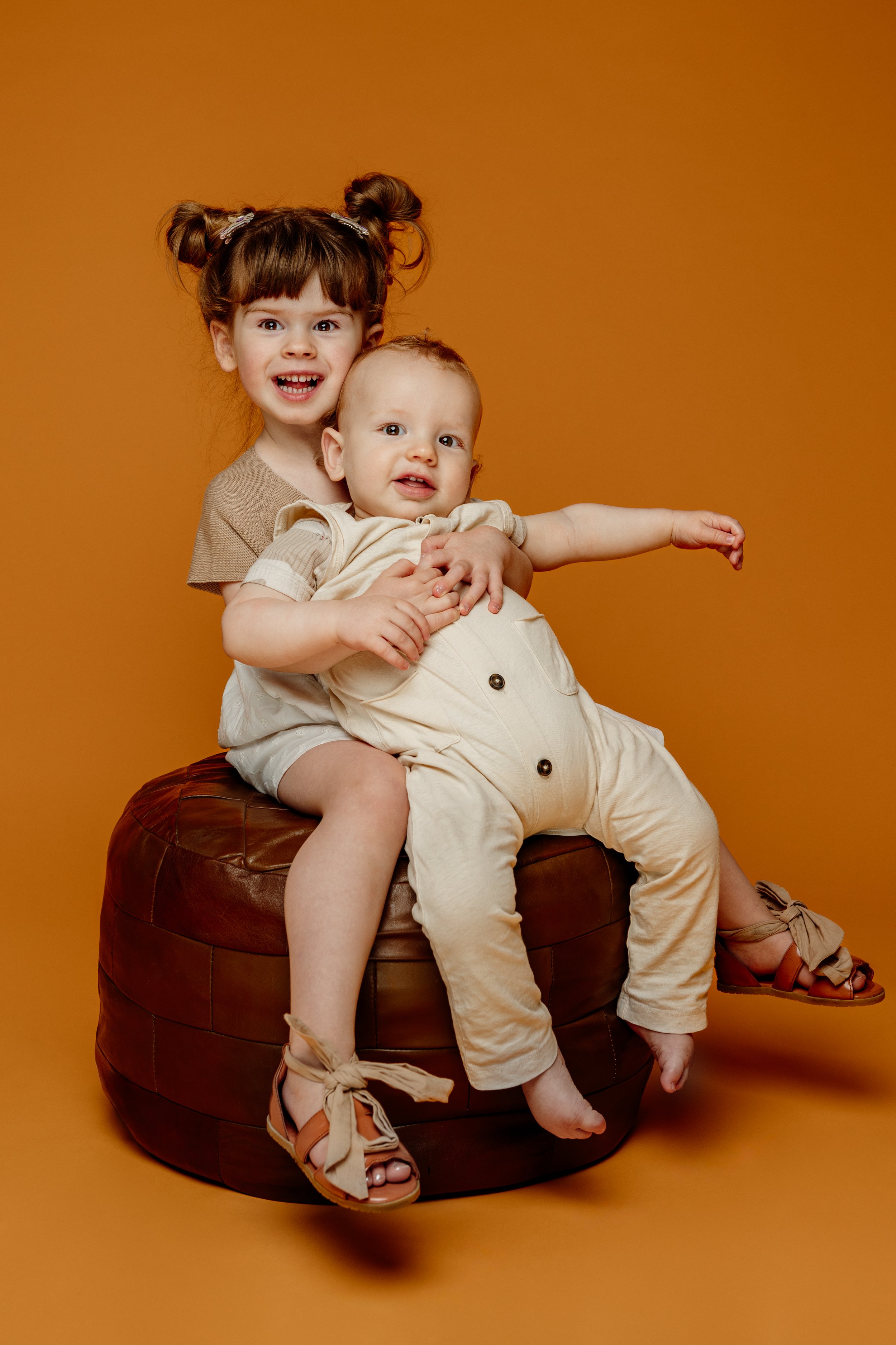 Twee jonge kinderen, een meisje en een jongen, zitten samen op een ronde, bruine zitbank tegen een oranje achtergrond. Het meisje heeft bruin haar in twee bunnen en lacht naar de camera. De jongen draagt een beige outfit en strekt zijn arm uit.
