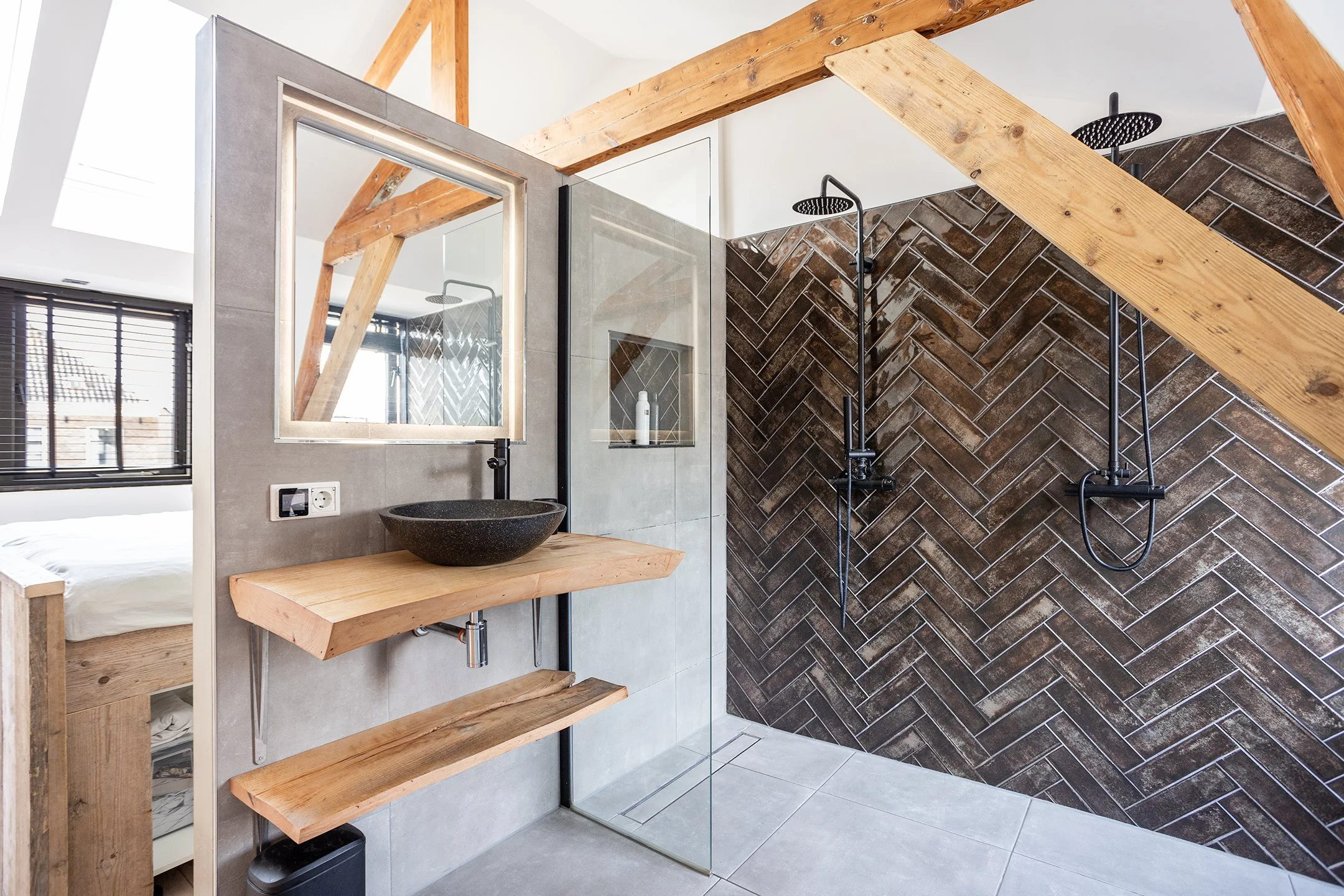 Een moderne badkamer met een inloopdouche en een houten wand. De douche heeft twee regendouches tegen een muur met een visgraatpatroon in donkere tegels. Er is een stenen wasbak boven een houten plank en een spiegel erboven. Een raam met zwarte kozij