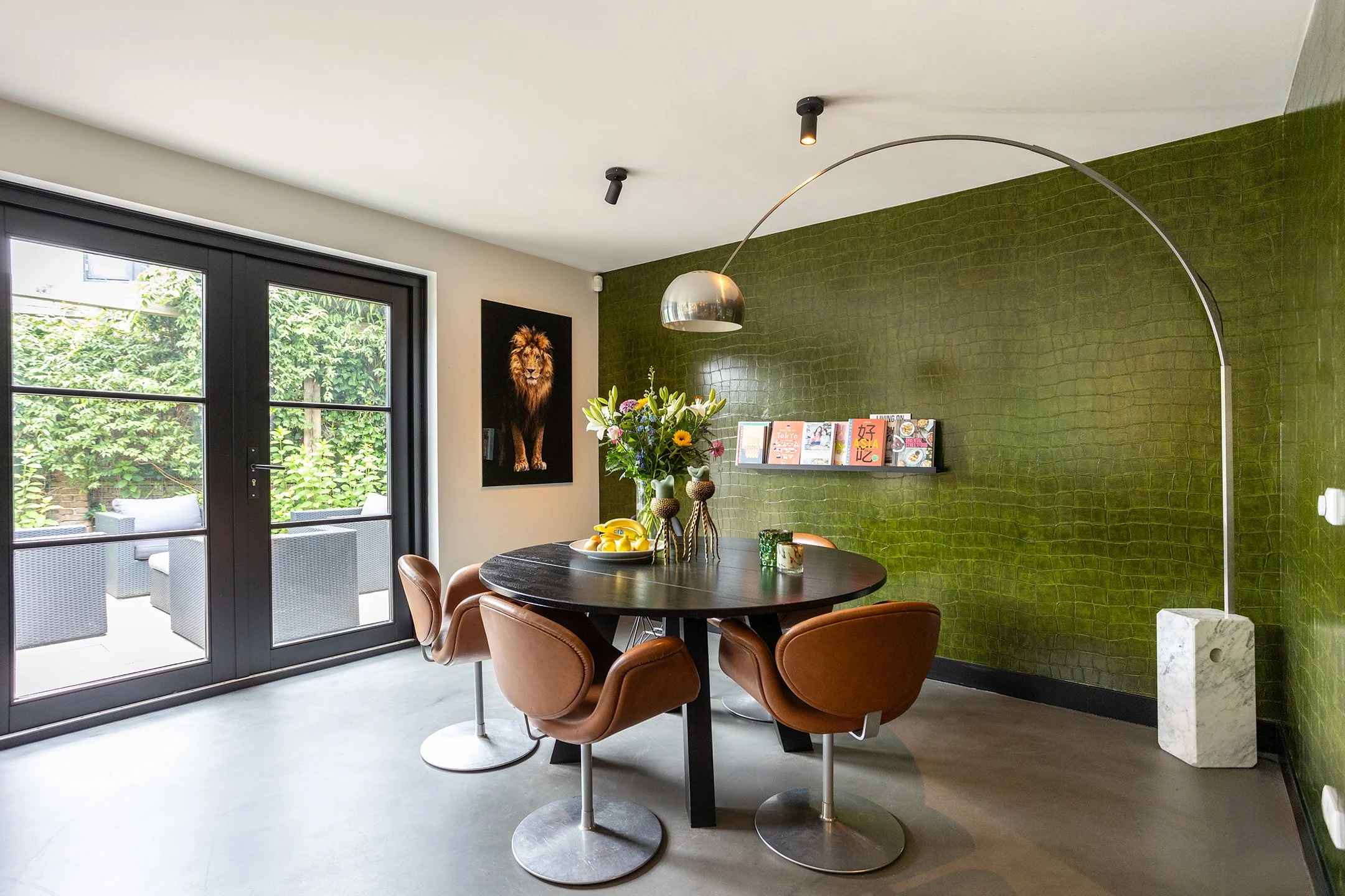 Een moderne eetkamer met een ronde zwarte tafel, zes bruin leren stoelen, een bloemenboeket, een muurschildering van een leeuw, een hanglamp en een groene beklede muur, met grote glazen deuren die uit open naar een balkon met tuinmeubilair leiden.