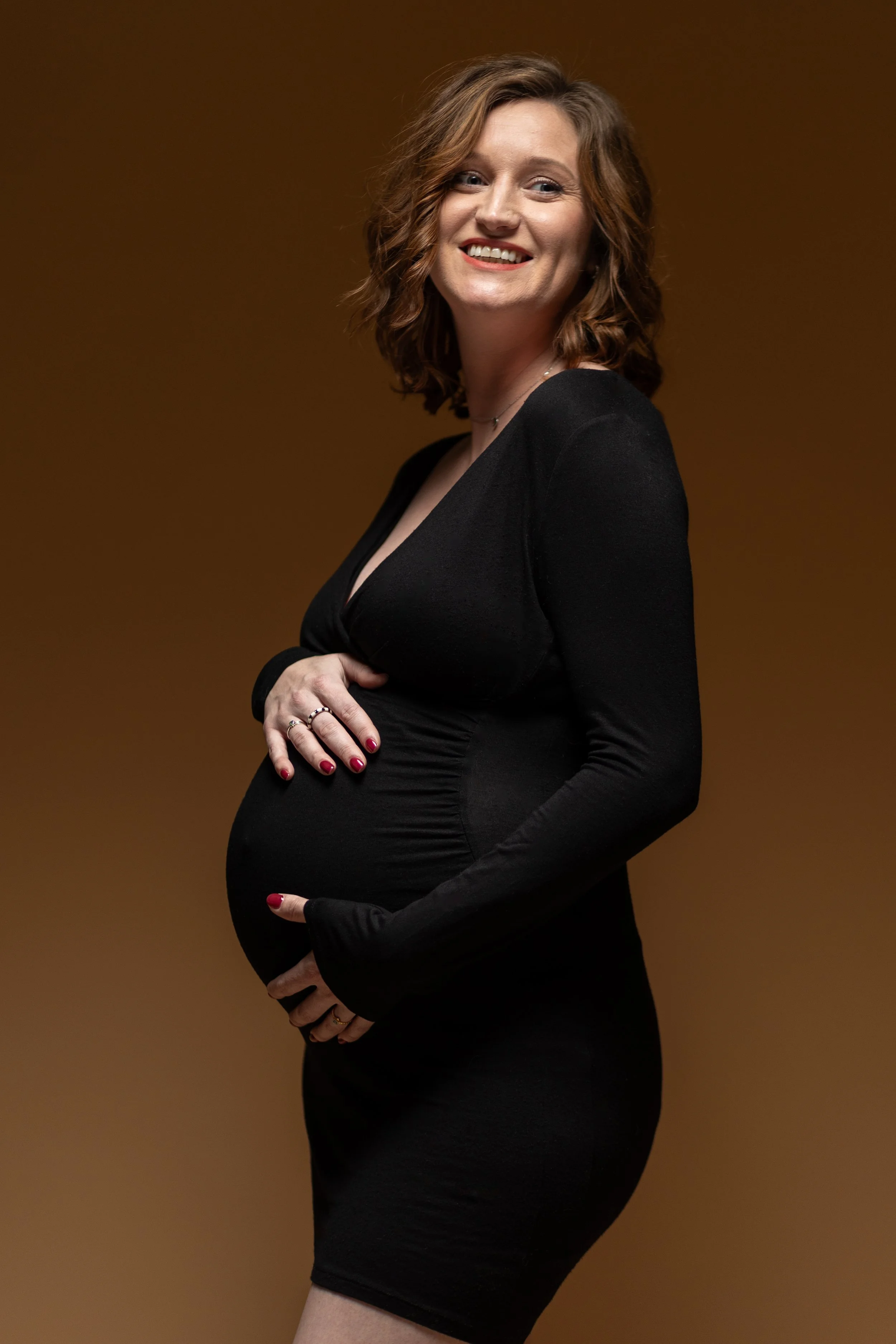 Portret van een zwangere vrouw met rood haar die lacht, gekleed in een zwarte jurk, op een beige achtergrond.