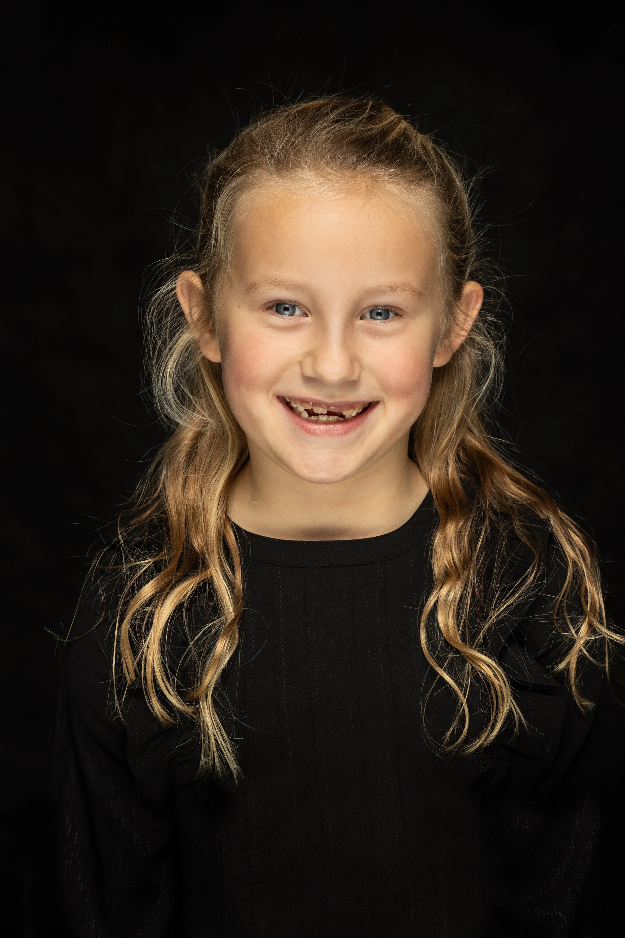 Portret van een lachend meisje met lang, golvend blond haar en blauwe ogen, gekleed in een zwarte top, tegen een zwarte achtergrond.