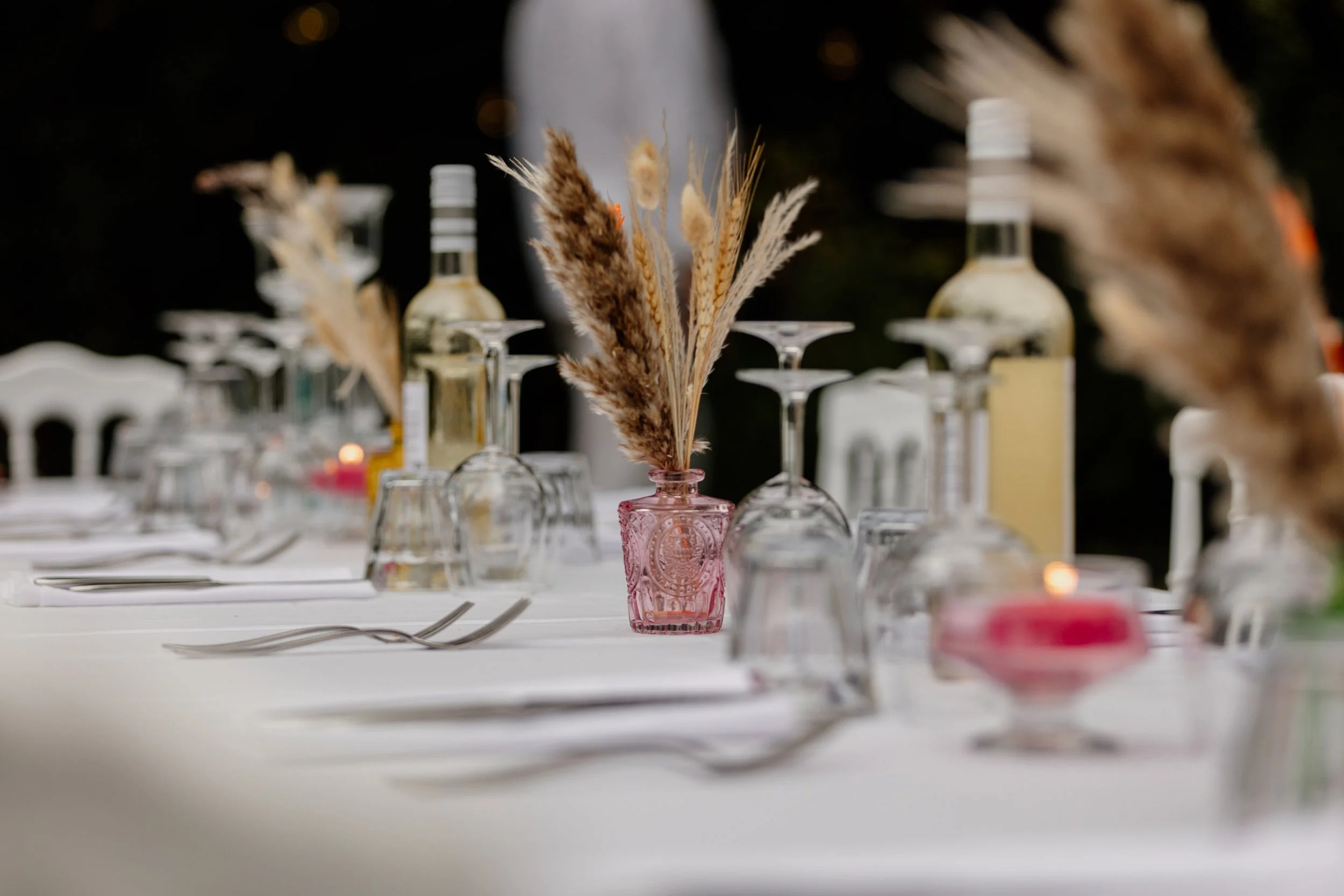Decoratieve tafelsetting met gedroogde bloemen in een roze glas, omgeven door wijn- en waterglazen, bestek en kaarsen, op een witte tafel tijdens een avondfeest.