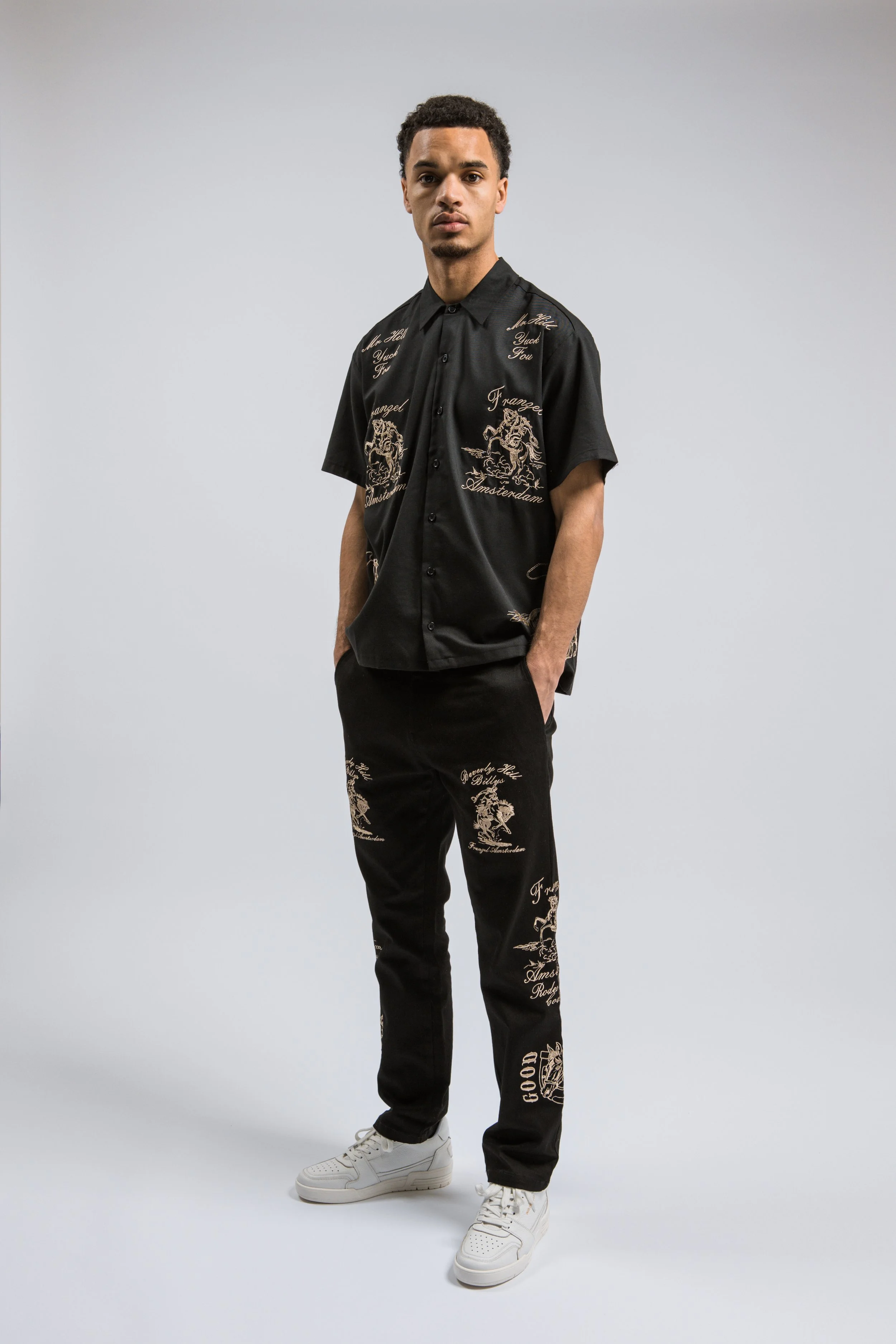 Een jonge man in een zwart gestreept shirt en broek met opvallende goudkleurige prints en woorden, gedragen op een neutrale grijze achtergrond.