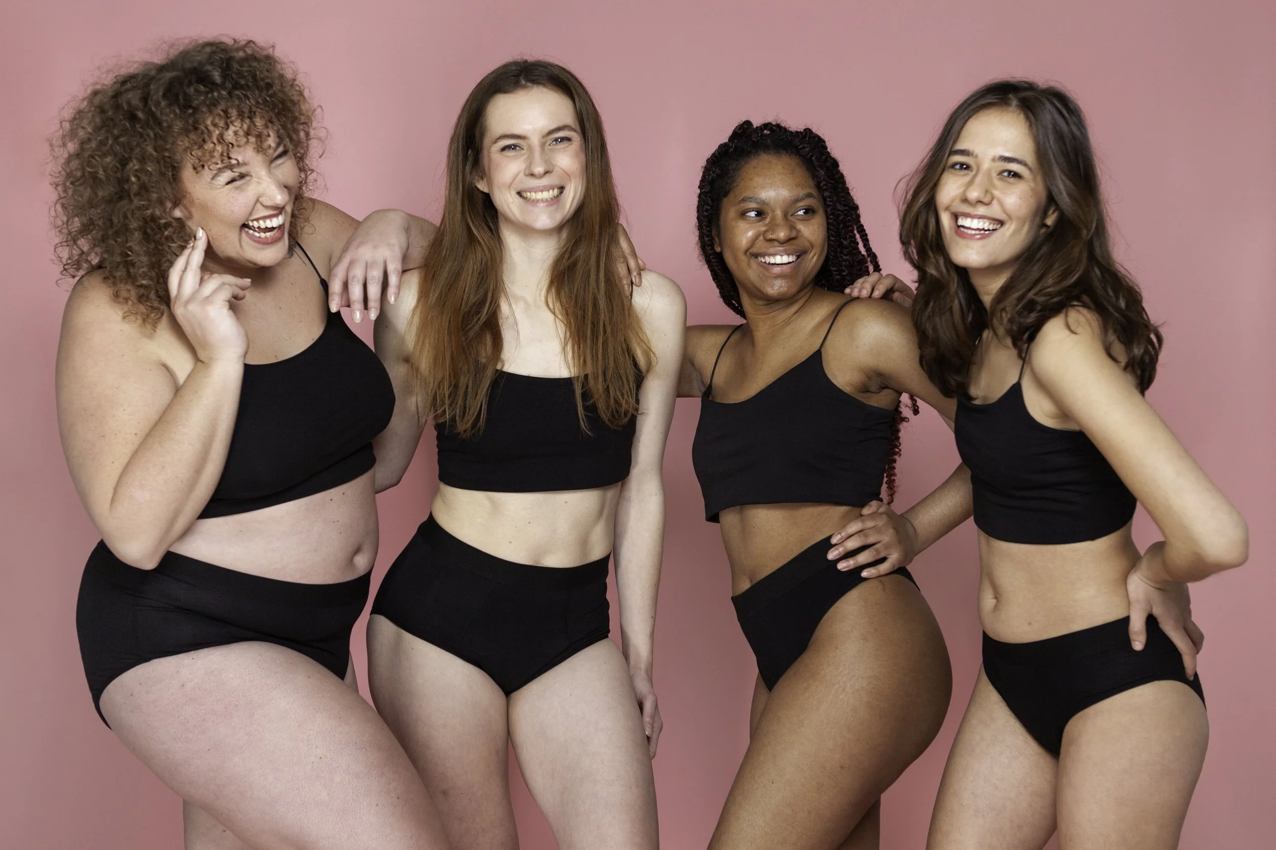 Vier jonge vrouwen lachen en poseren samen tegen een roze achtergrond, gekleed in zwarte sportkleding.