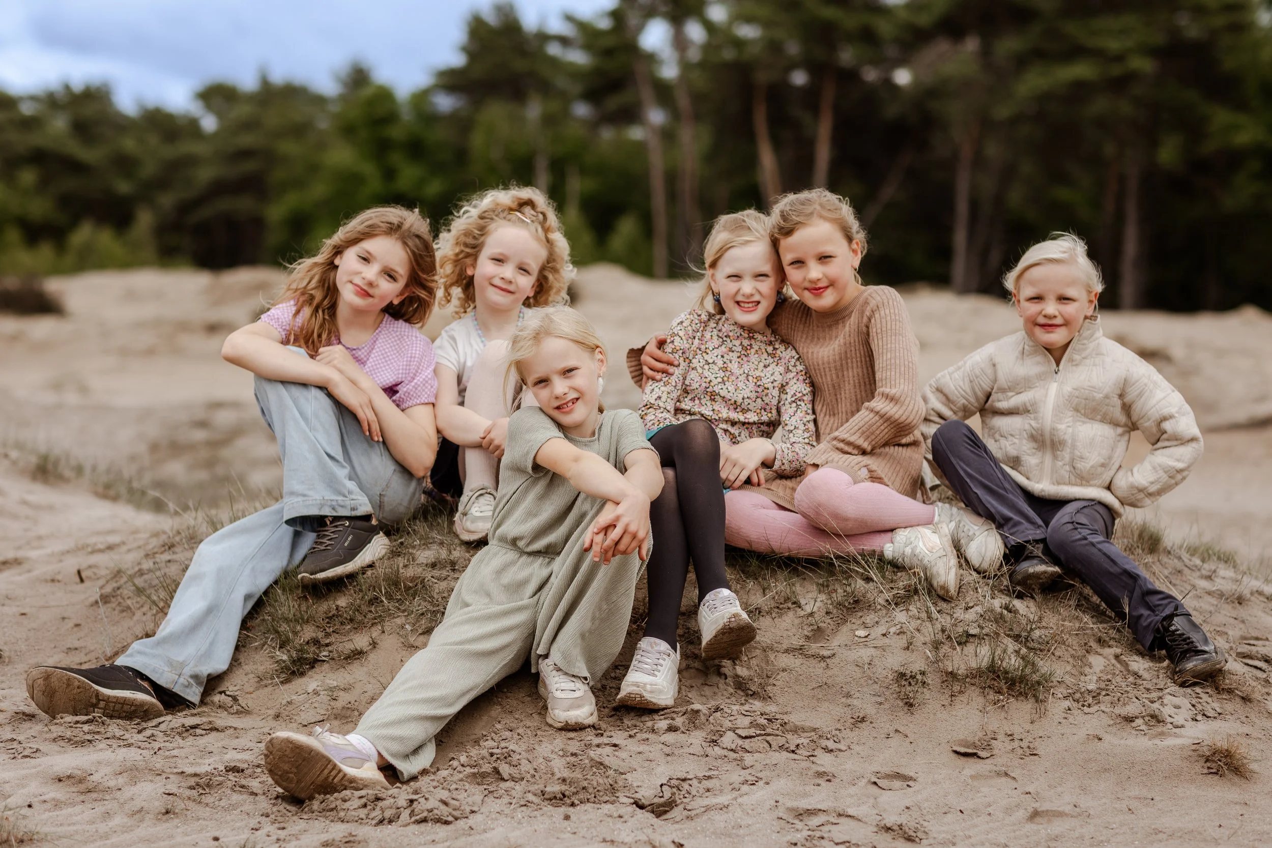 Zeven jonge meisjes zitten en liggen op een zandheuvel in een bos, glimlachend naar de camera.