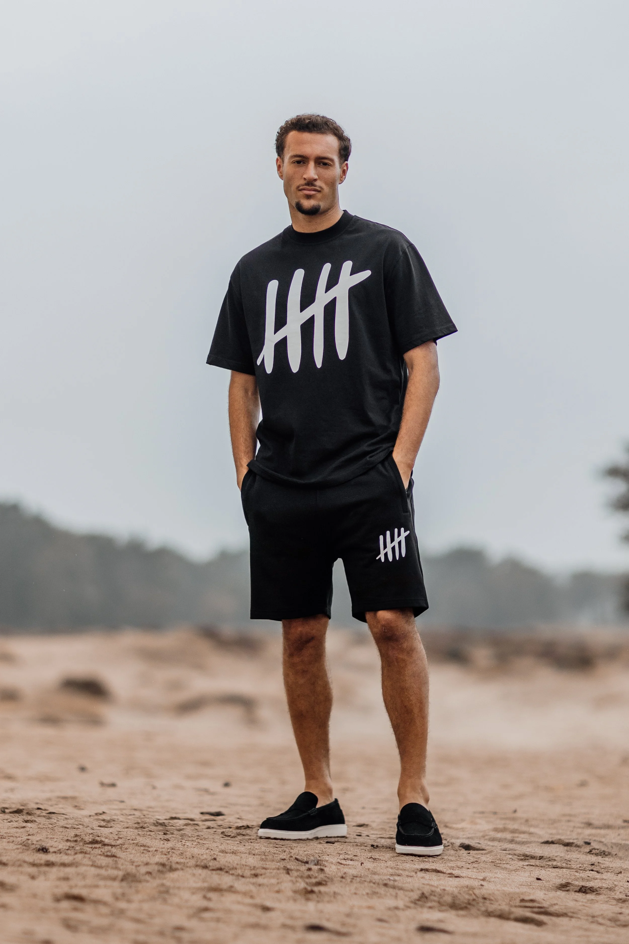 Man staat op een zandstrand, gekleed in een zwarte T-shirt en shorts met logo's, met handen in zakken, onder een bewolkte lucht.