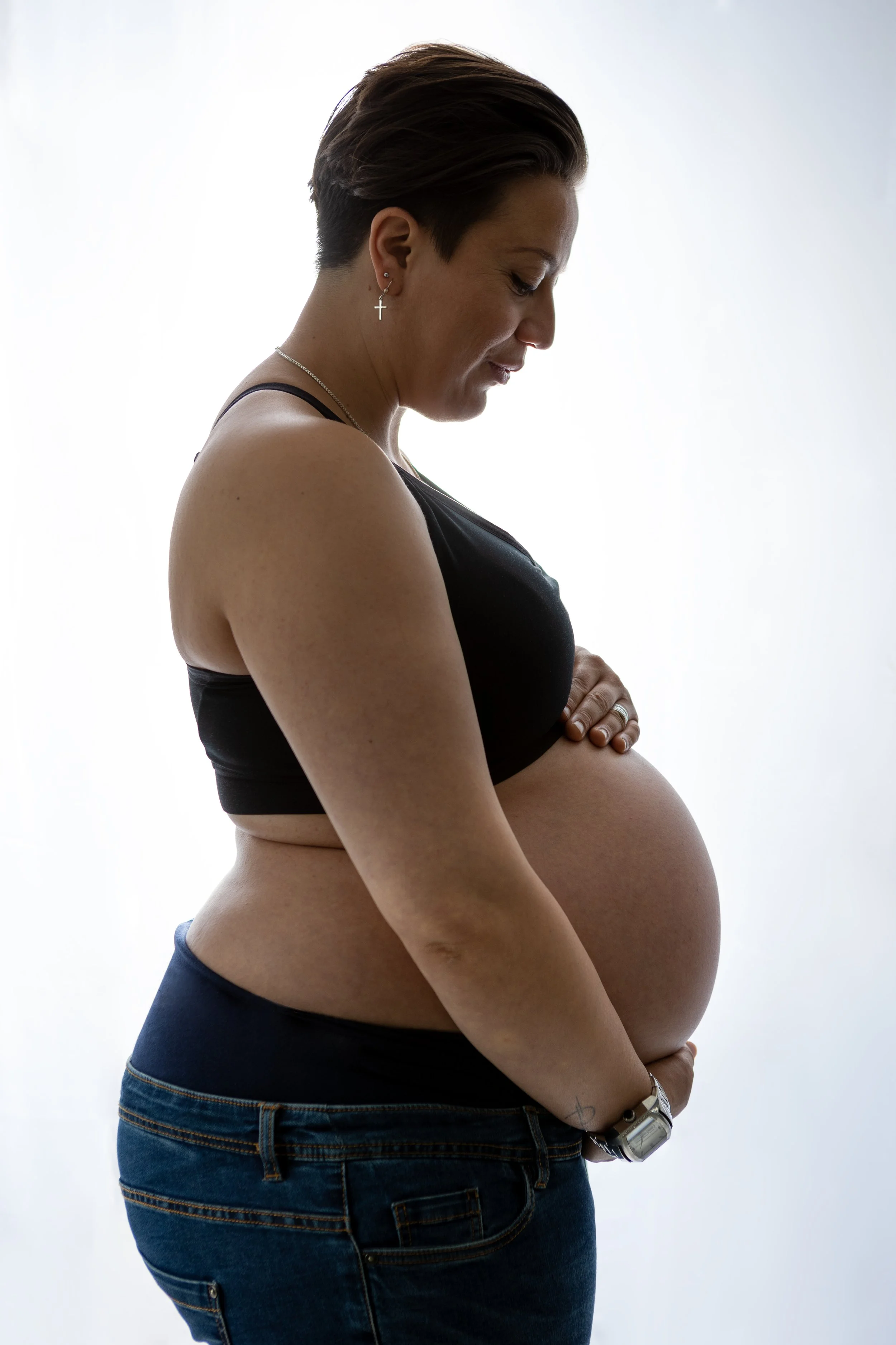 Zwangere vrouw met korte haar draagt een zwarte top en jeans, kijkt naar haar buik.