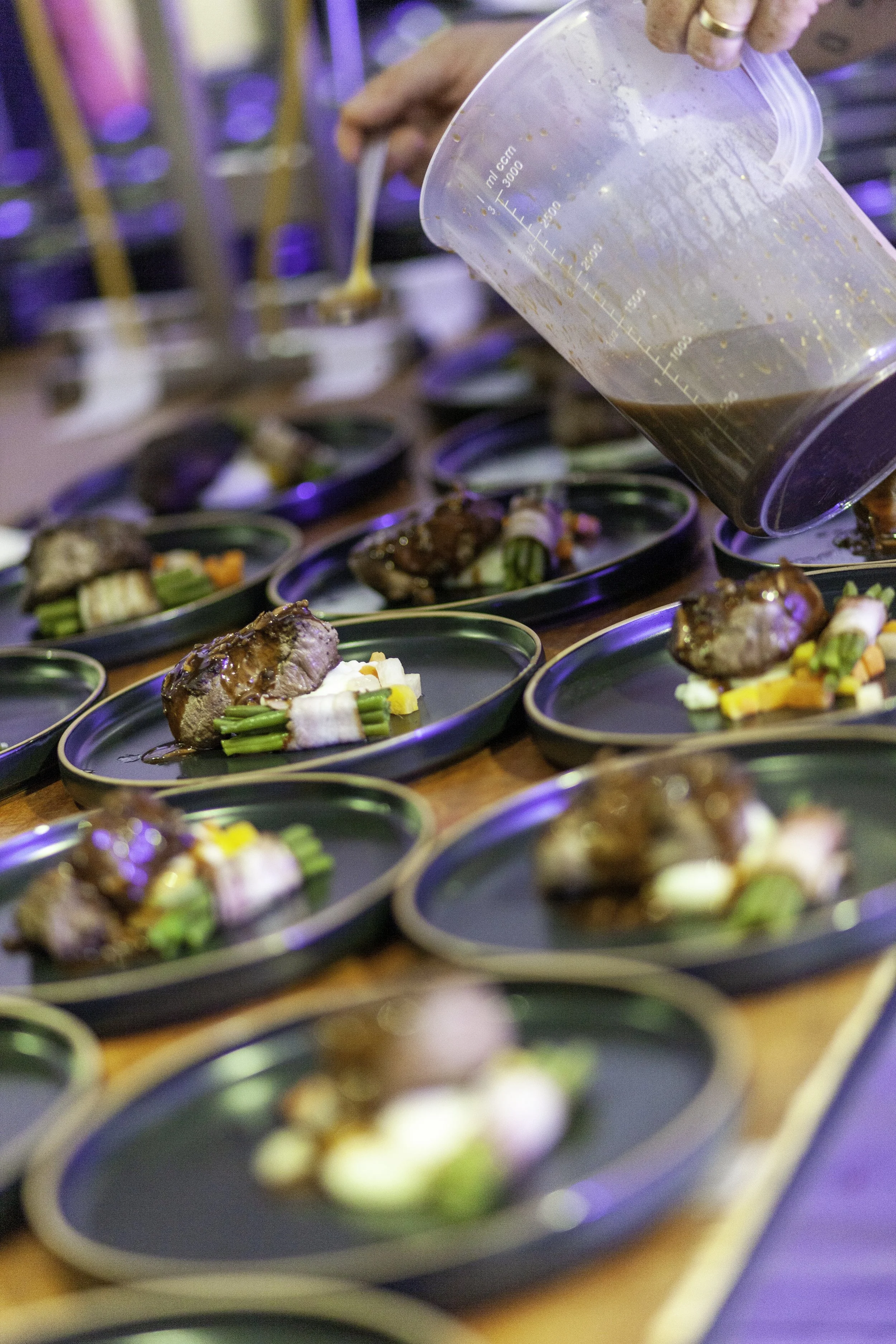 Een persoon schenkt saus over kleine borden met vlees, groenten en garnituren op een tafel.