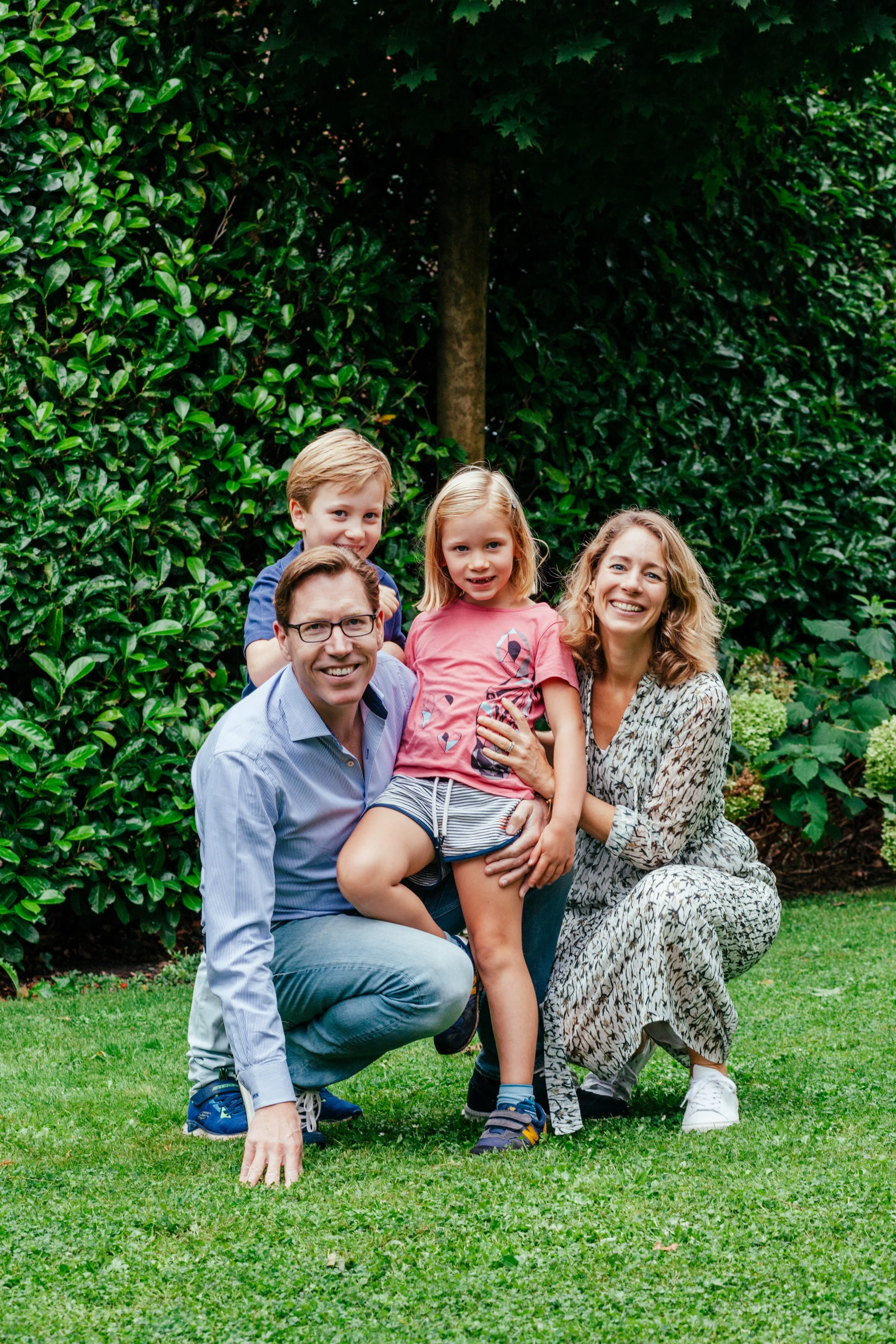 Een gezin van vier personen dat buiten op het gazon staat, omringd door groene heggen en struiken, lachend voor de camera.