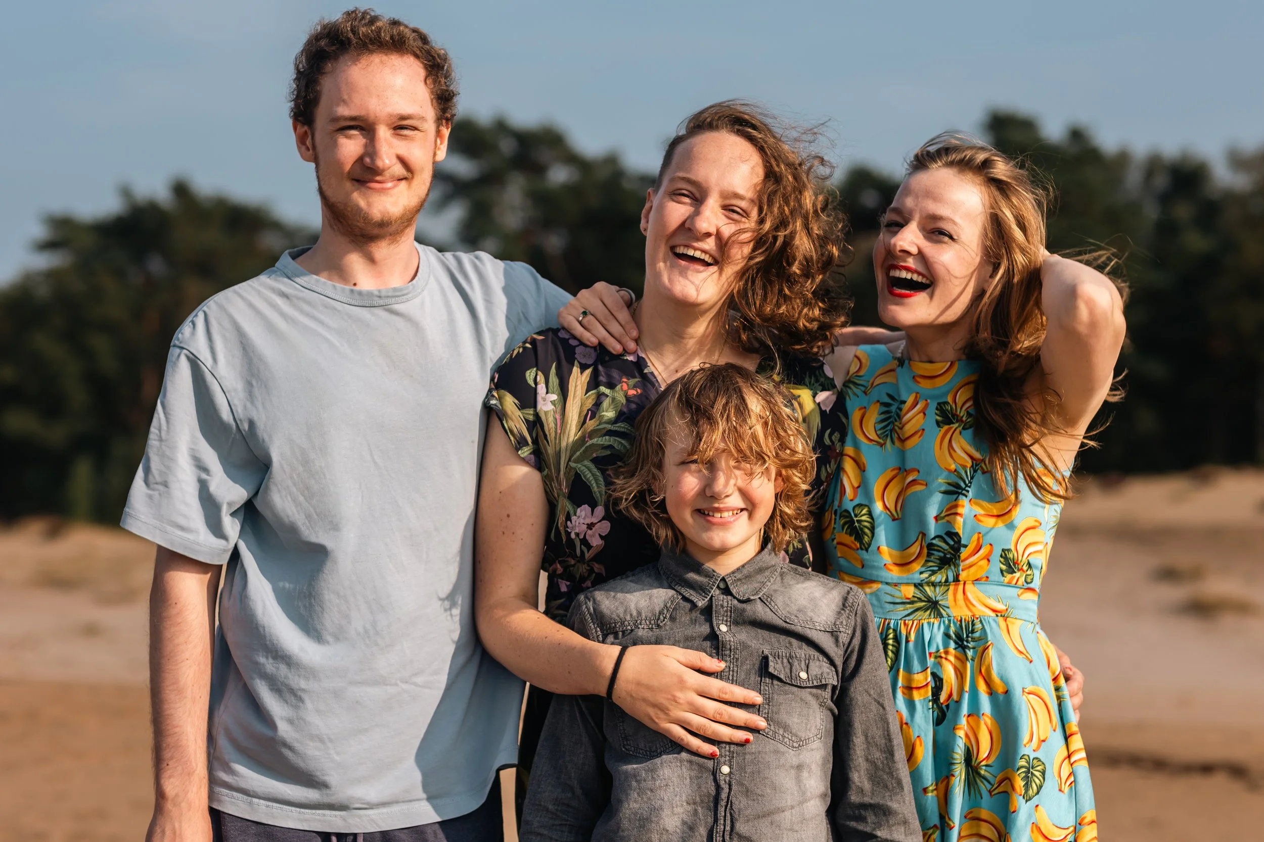 Vijf mensen lachen en poseren samen buiten op een dag met blauwe hemel en bomen op de achtergrond.