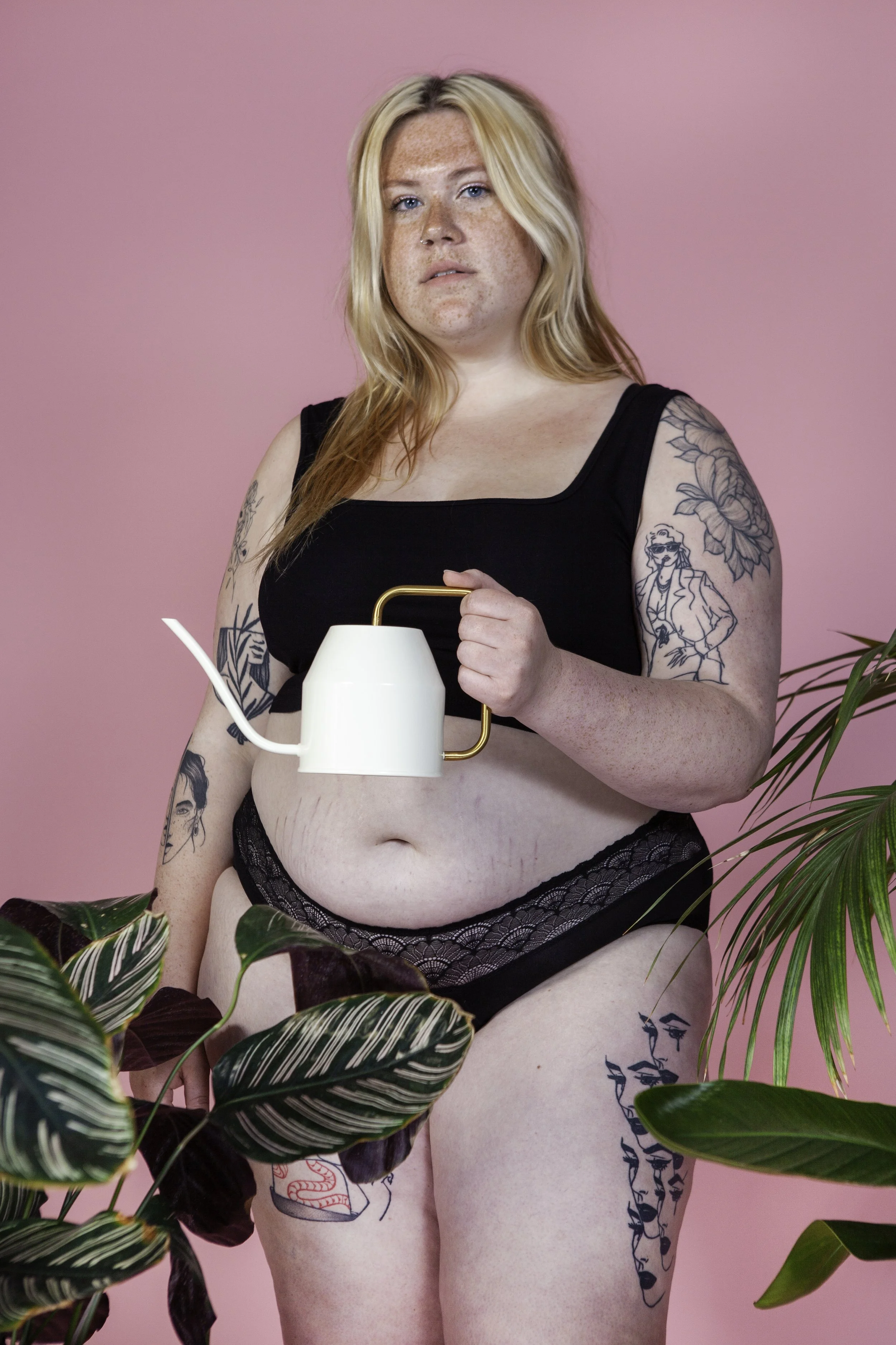 Een vrouw met tattoos draagt zwart lingerie en houdt een witte en goudkleurige waterkoker vast voor een fotoshoot, met een roze achtergrond en groene planten op de voorgrond.