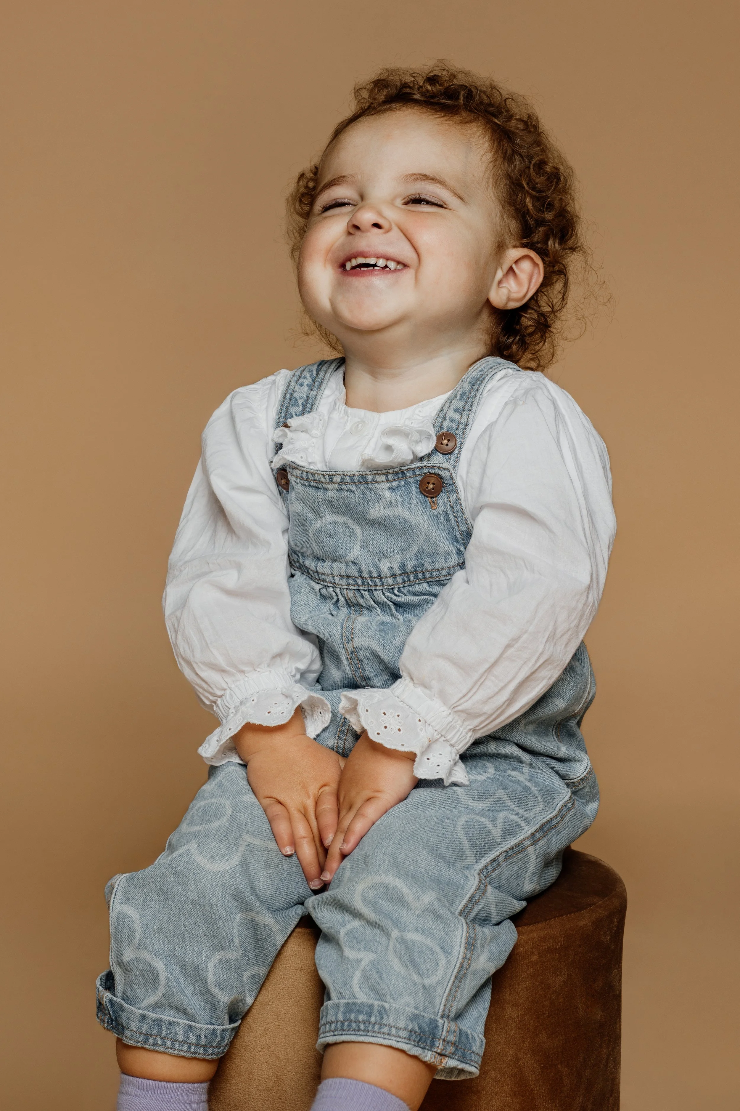 Een lachende jonge jongen met lichtbruin, krullend haar zit op een bruine kruk, gekleed in een witte blouse en blauwe denim overalls met patroon en details. De achtergrond is beige.