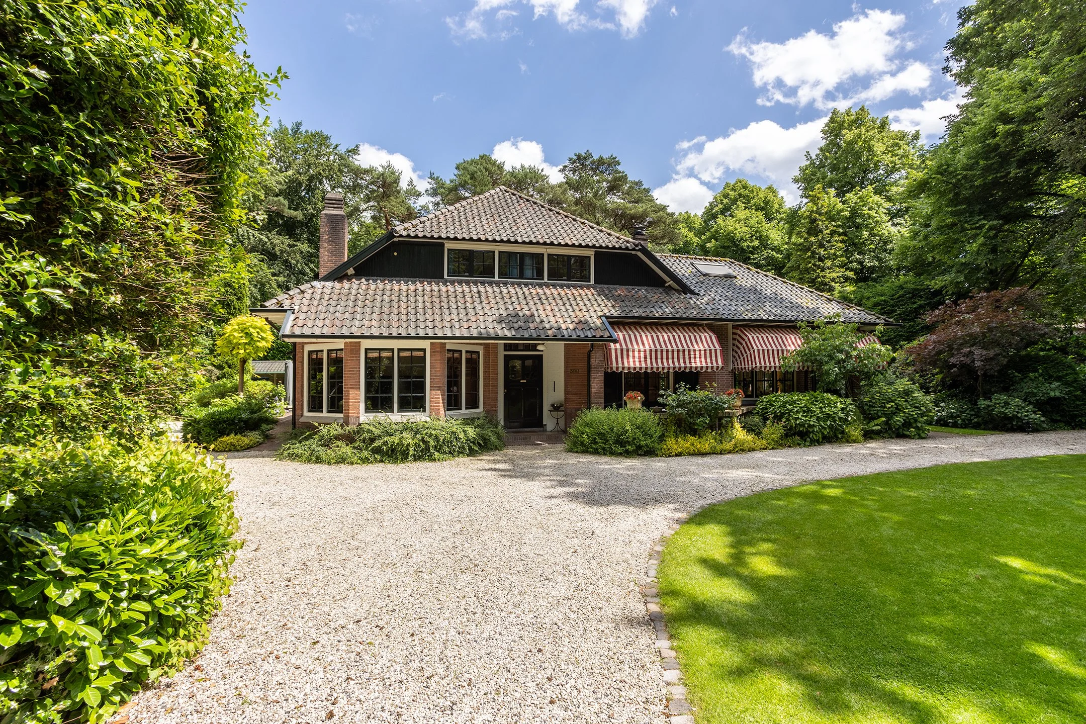 Gezicht op een huis met een grote tuin, omgeven door bomen en struiken, met een gravelpad voor de ingang.