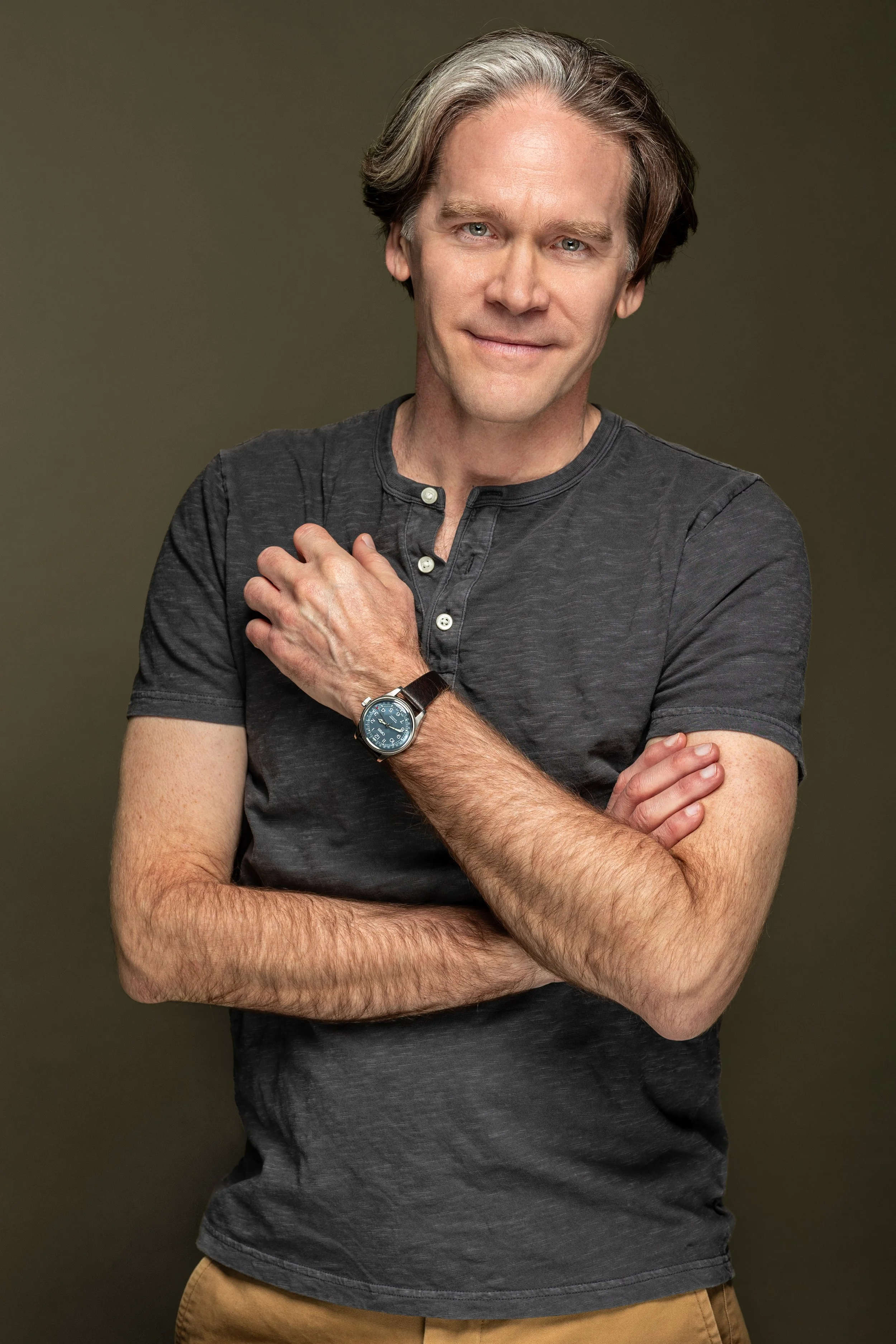 Man met grijze haar en blauwe ogen die een grijs T-shirt draagt en een horloge draagt, poseert tegen een effen achtergrond.