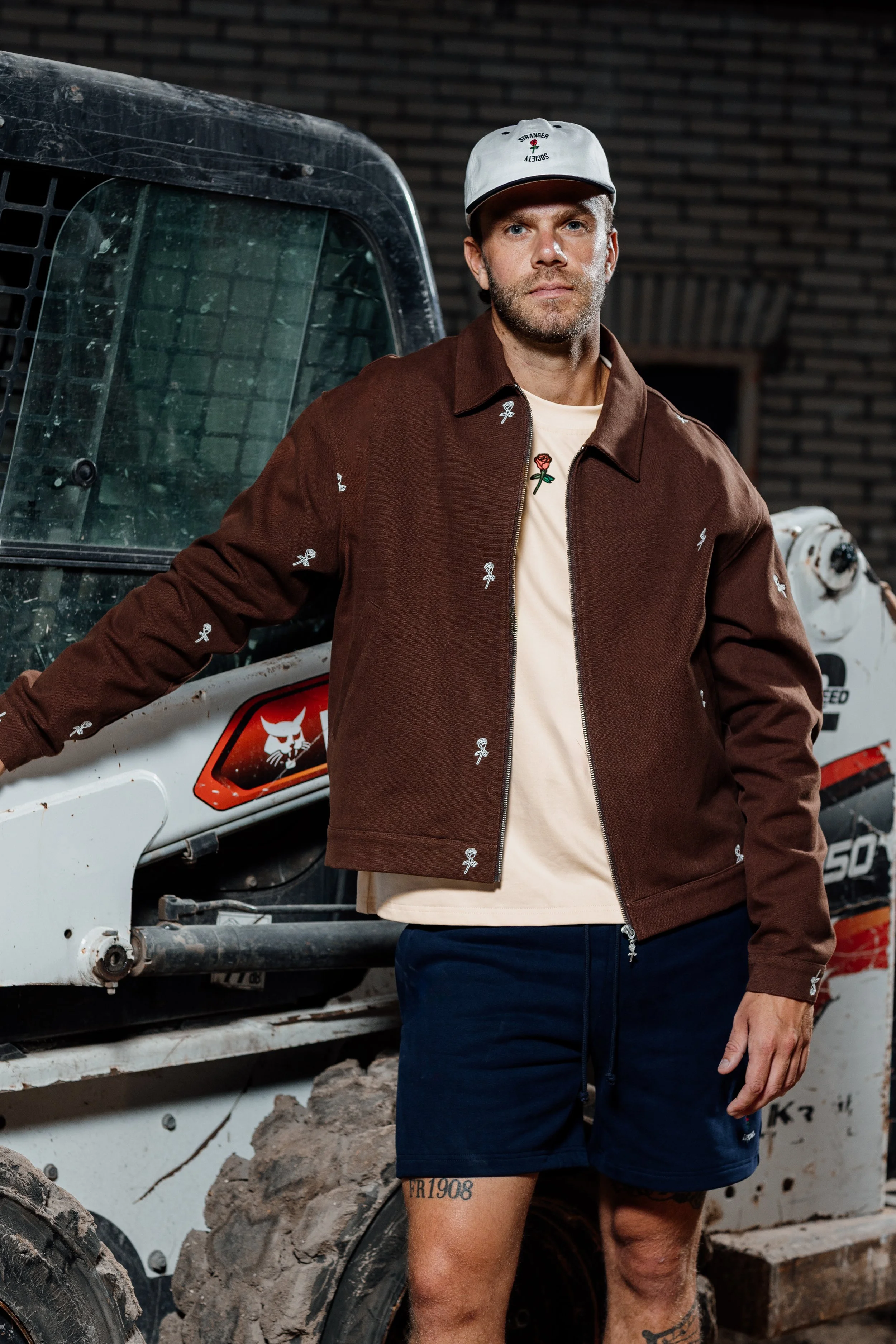 Een man met een pet en een bruine jas met kleine witte bloemmotieven, staand naast een bouwvoertuig in een industriële omgeving.