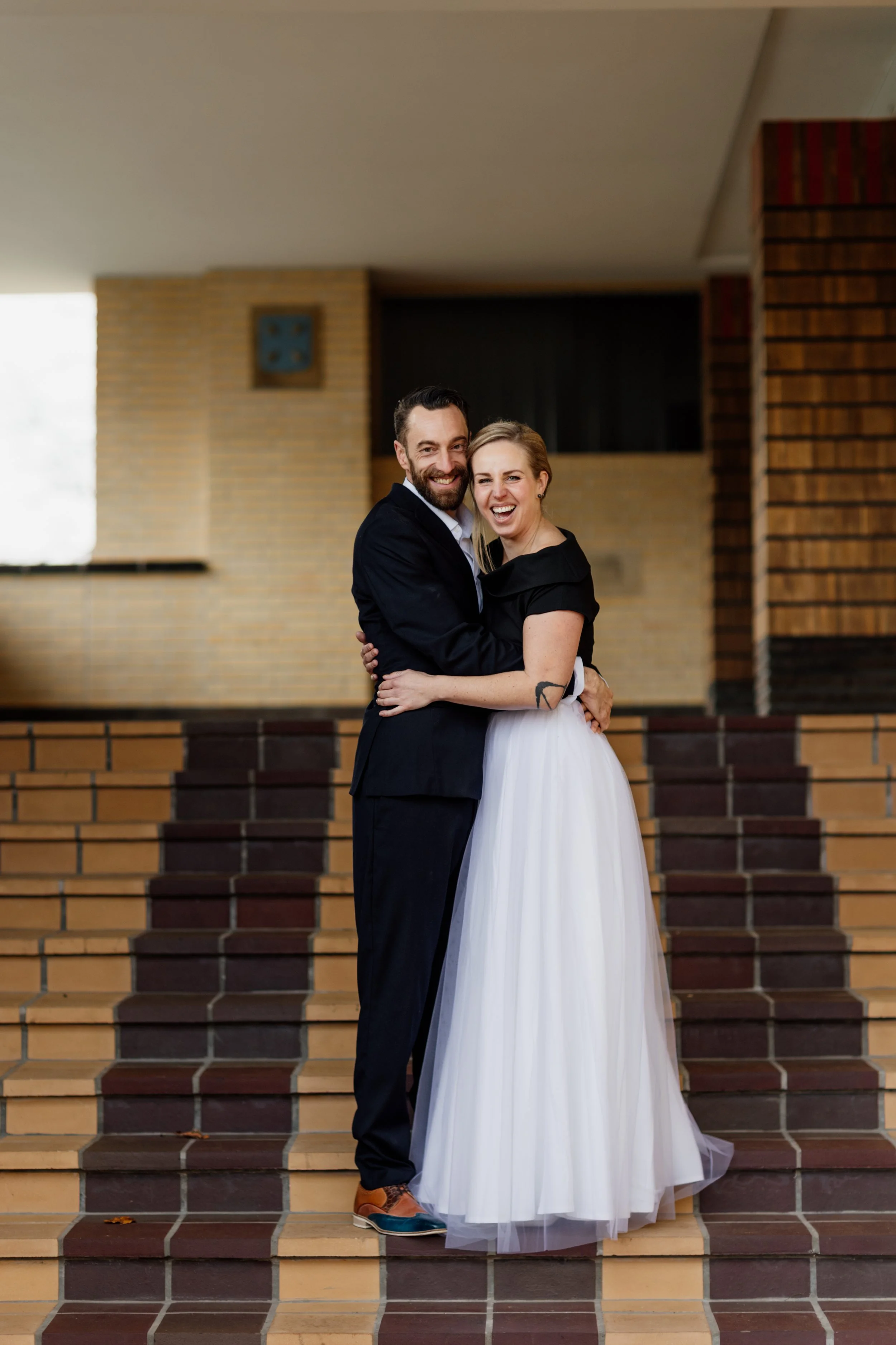 Een stel in huwelijkskleding, lachend en knuffelend op een trap in een binnenruimte, met bakstenen muren op de achtergrond.