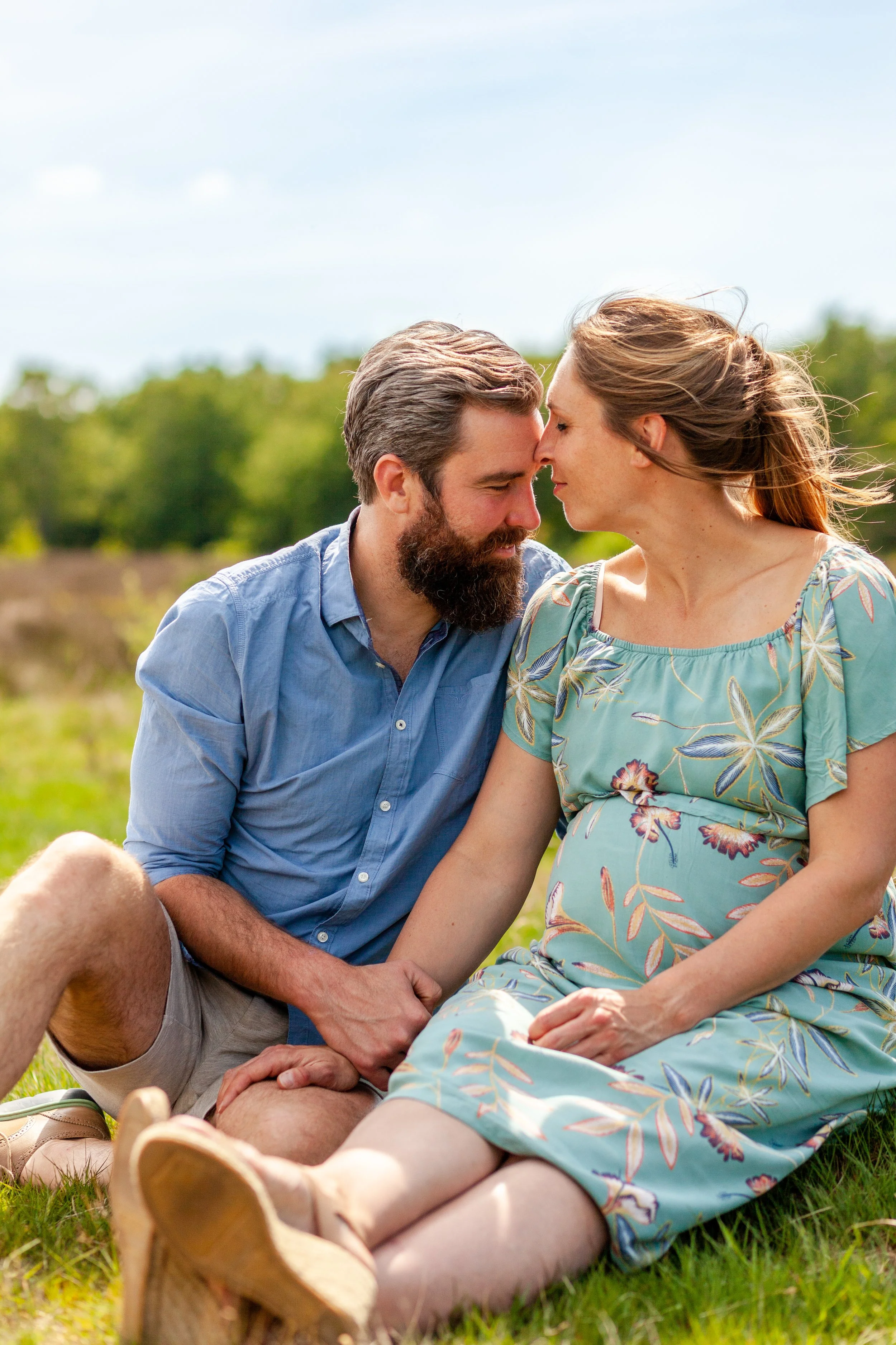 Een koppel zit samen in een grasveld, met hun voorhoofden tegen elkaar, onder een blauwe lucht. De man heeft een baard en draagt een lichtblauwe blouse, de vrouw draagt een mintgroene jurk met bloemenpatroon.