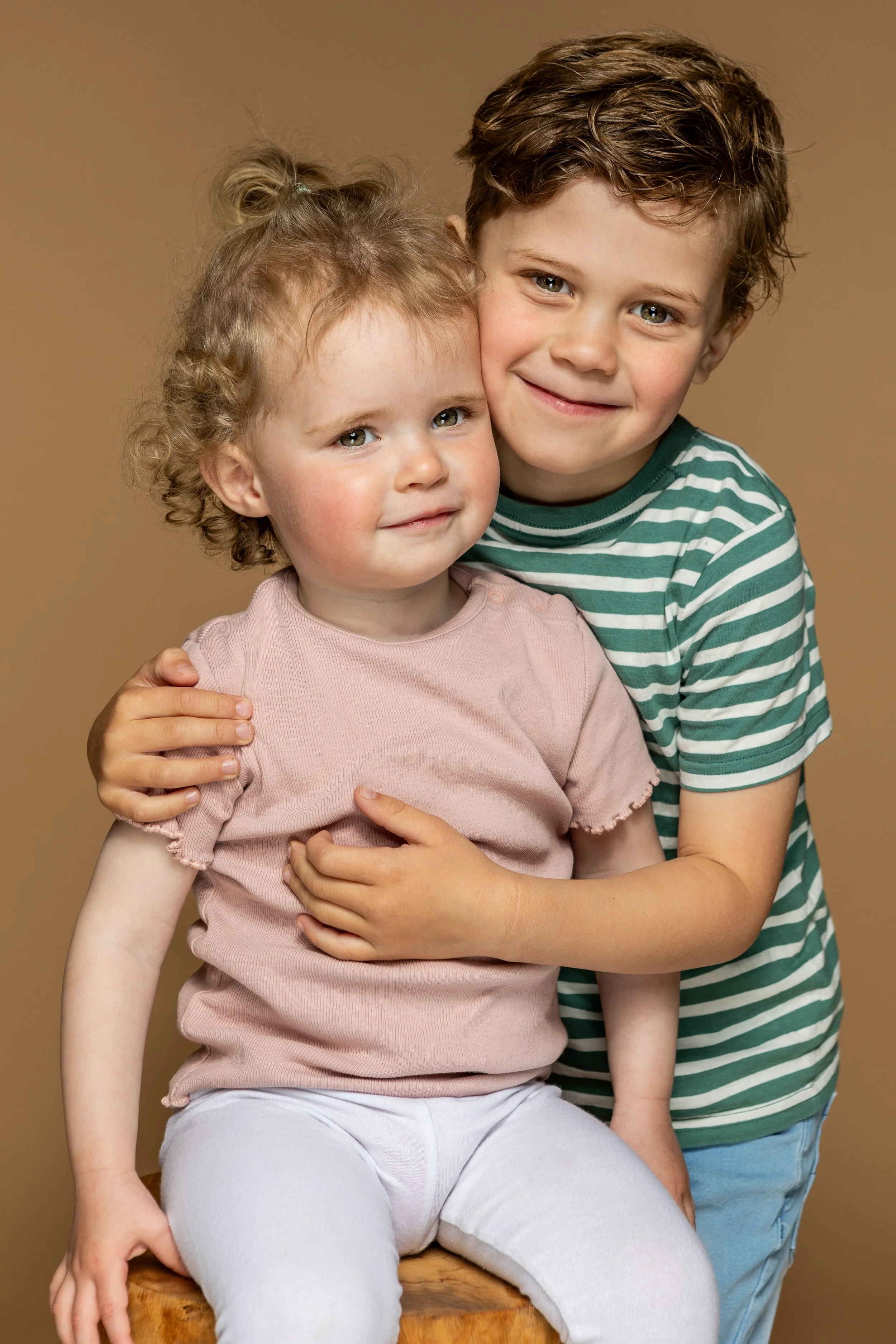 Twee kinderen, een meisje en een jongen, knuffelen elkaar en glimlachen, met een effen bruine achtergrond.