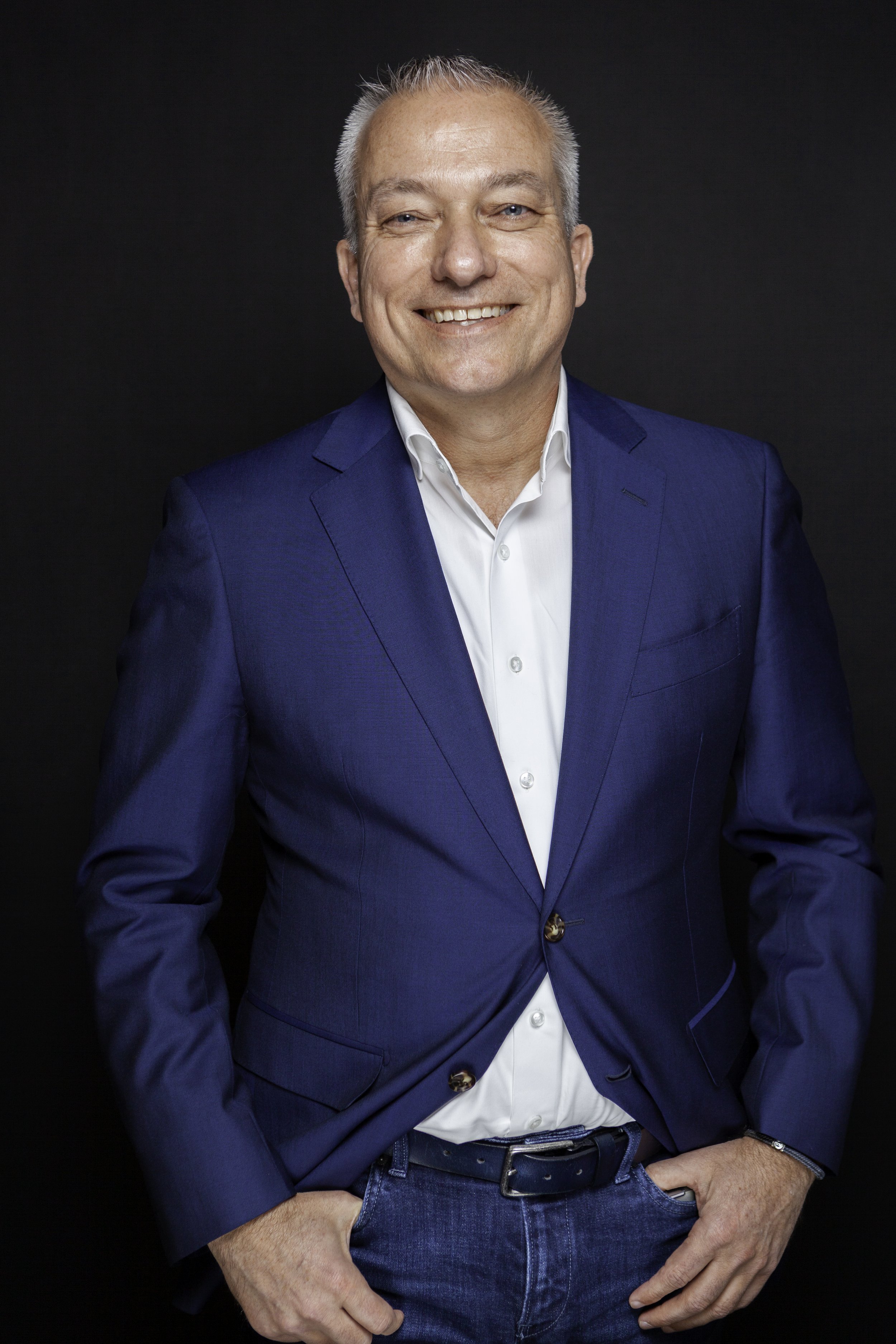 Een man met kort, grijs haar, lachend, gekleed in een blauwe blazer en witte overhemd, poseert voor een zwarte achtergrond.