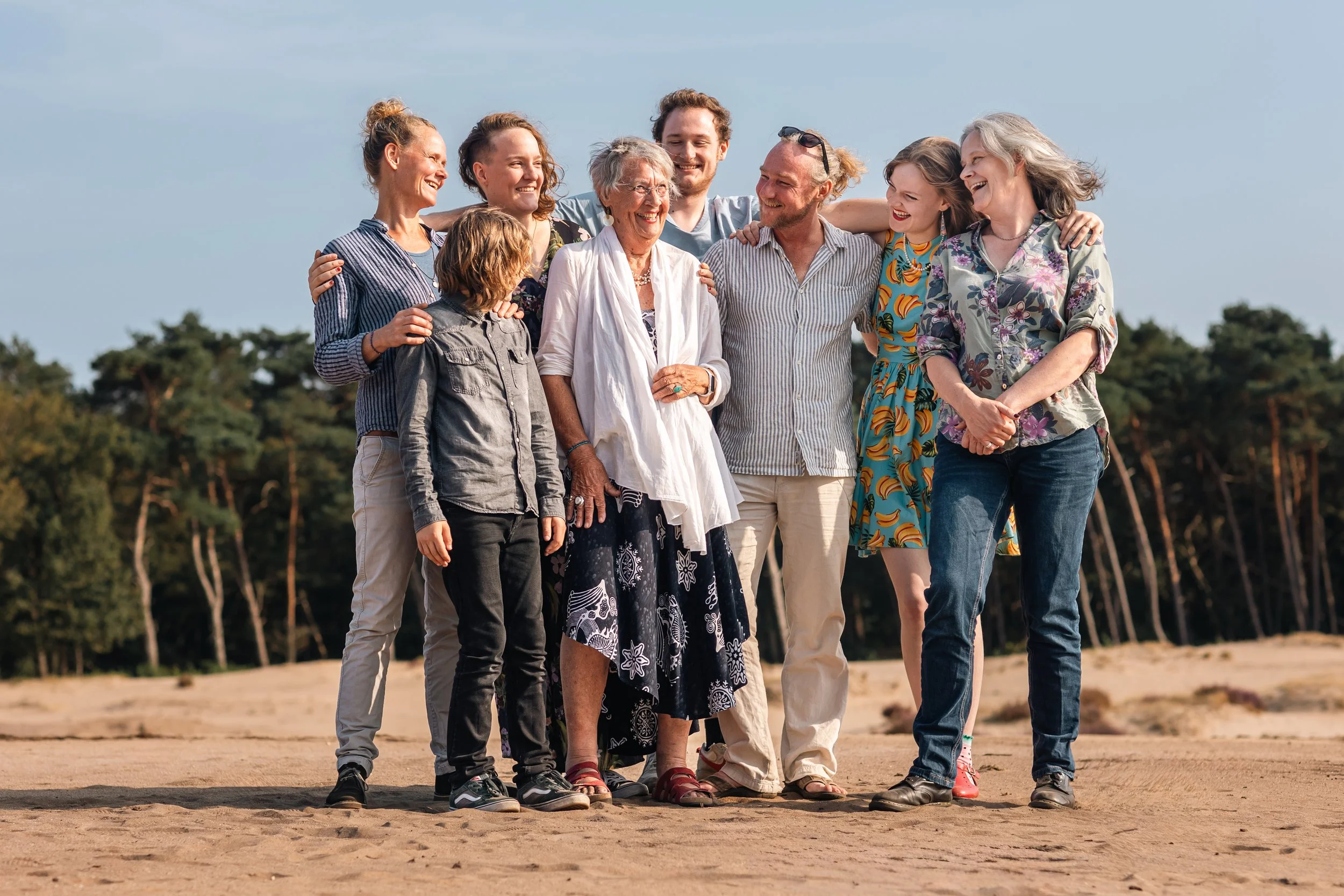 Een groep mensen, bestaande uit zeven volwassenen en een kind, die samen op een zandvlak stappen, lachend en in een kring vorm. Op de achtergrond bomen en een heldere hemel.