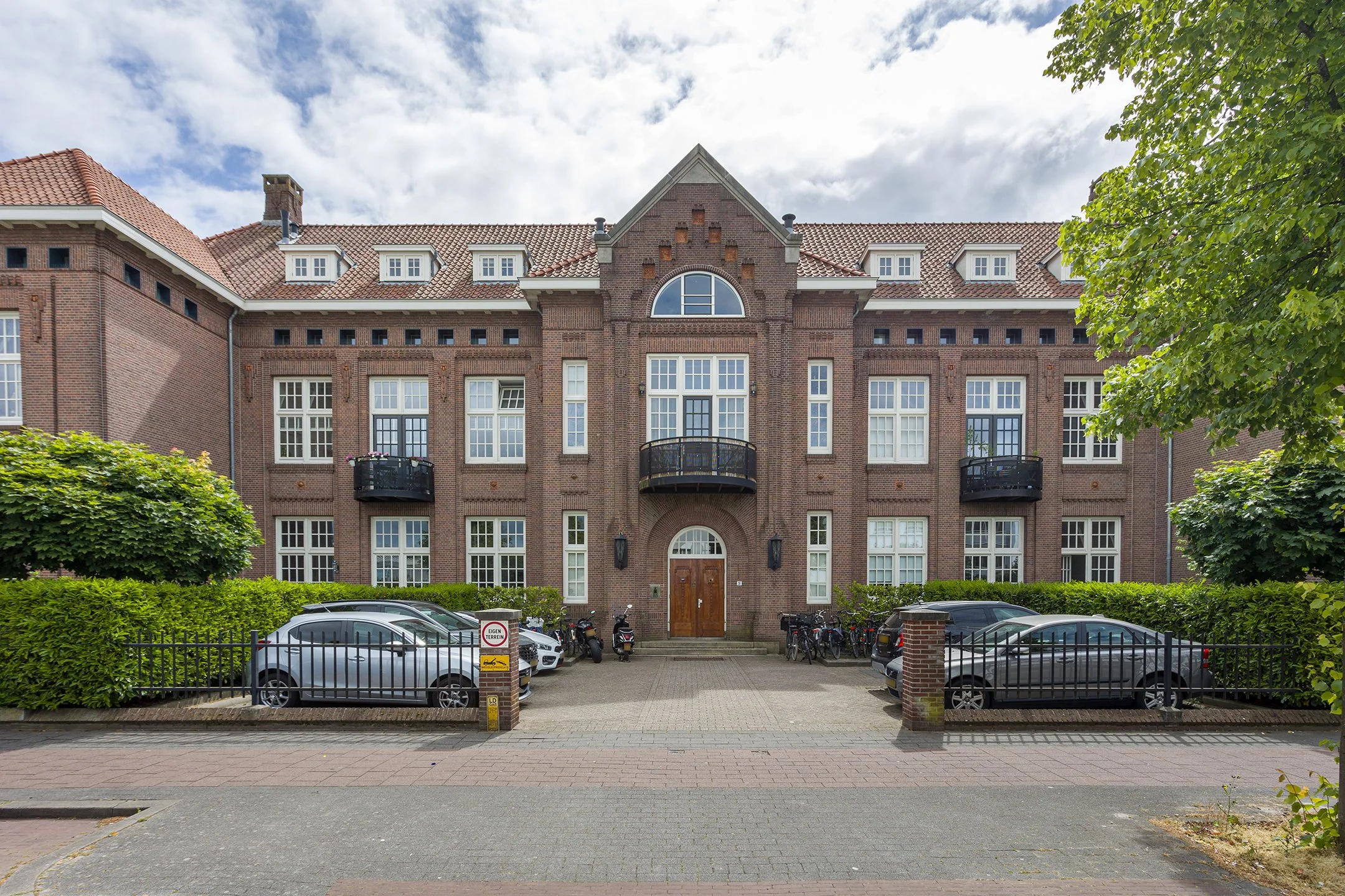 Een groot, oud bakstenen appartementencomplex met meerdere verdiepingen, balkonnetjes, witte ramen en een centrale houten deur. Er staan auto's en motoren geparkeerd in de voortuin, omgeven door groene struiken en bomen, onder een deels bewolkte heme