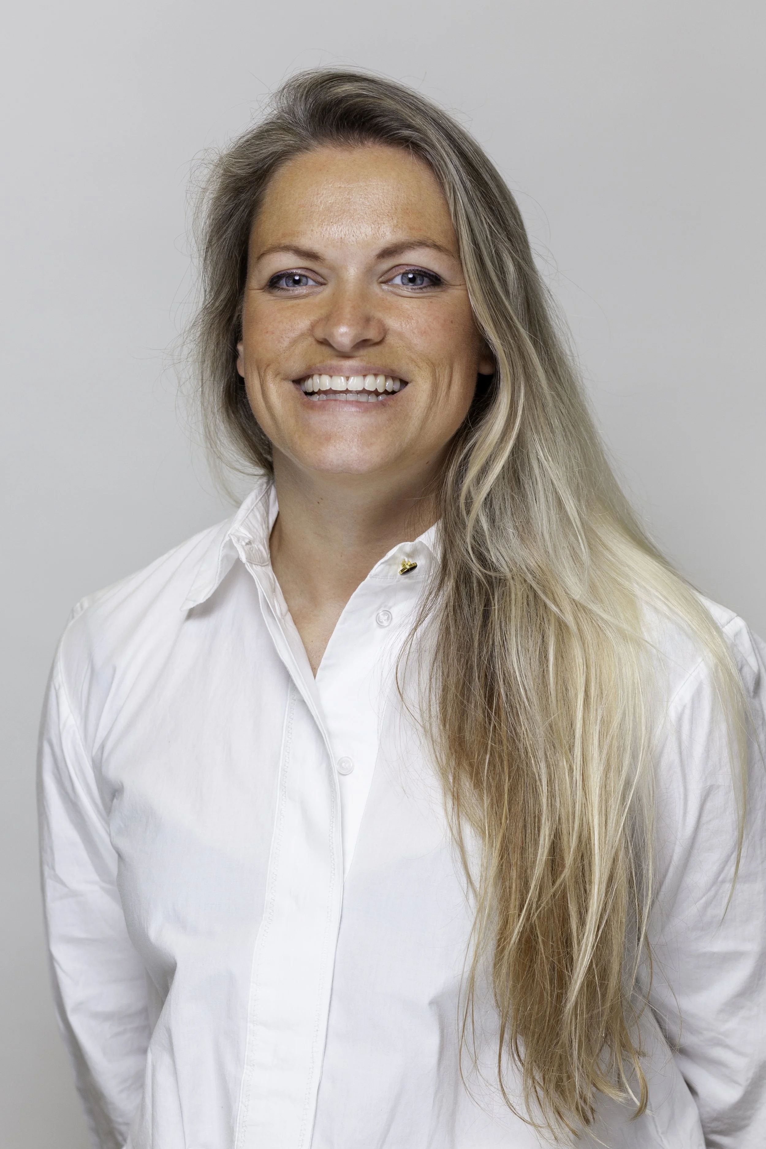 Een lachende vrouw met lang blond haar, gekleed in een witte blouse, voor een lichte achtergrond.