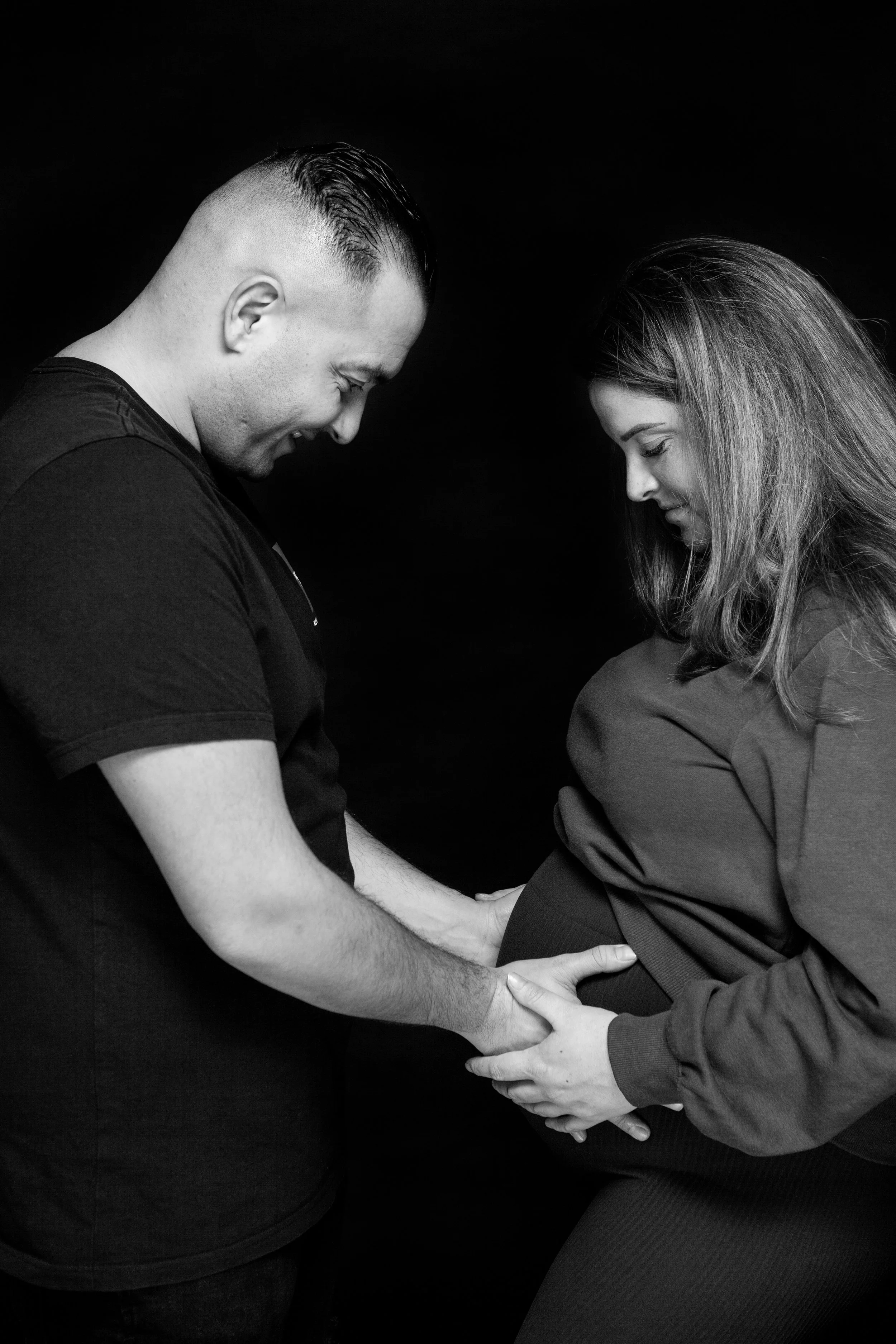 Zwart-wit foto van een man die een zwangere vrouw helpt met haar buik vast te houden, beiden lachen en kijken naar elkaar, donkere achtergrond.