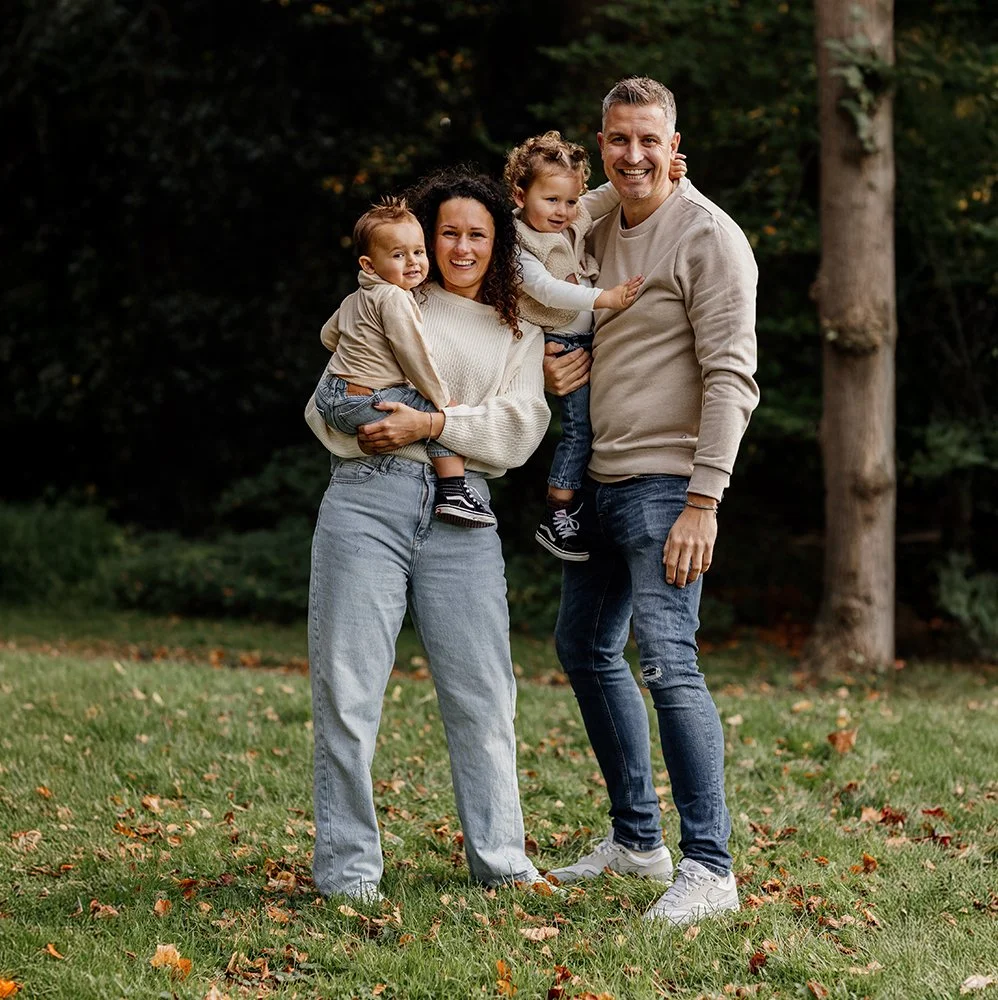 Gezinnetje van vier mensen, twee ouders en twee jonge kinderen, staan samen buiten in een park met bomen op de achtergrond. Ze lachen en knuffelen elkaar, gekleed in casual herfstkleding.