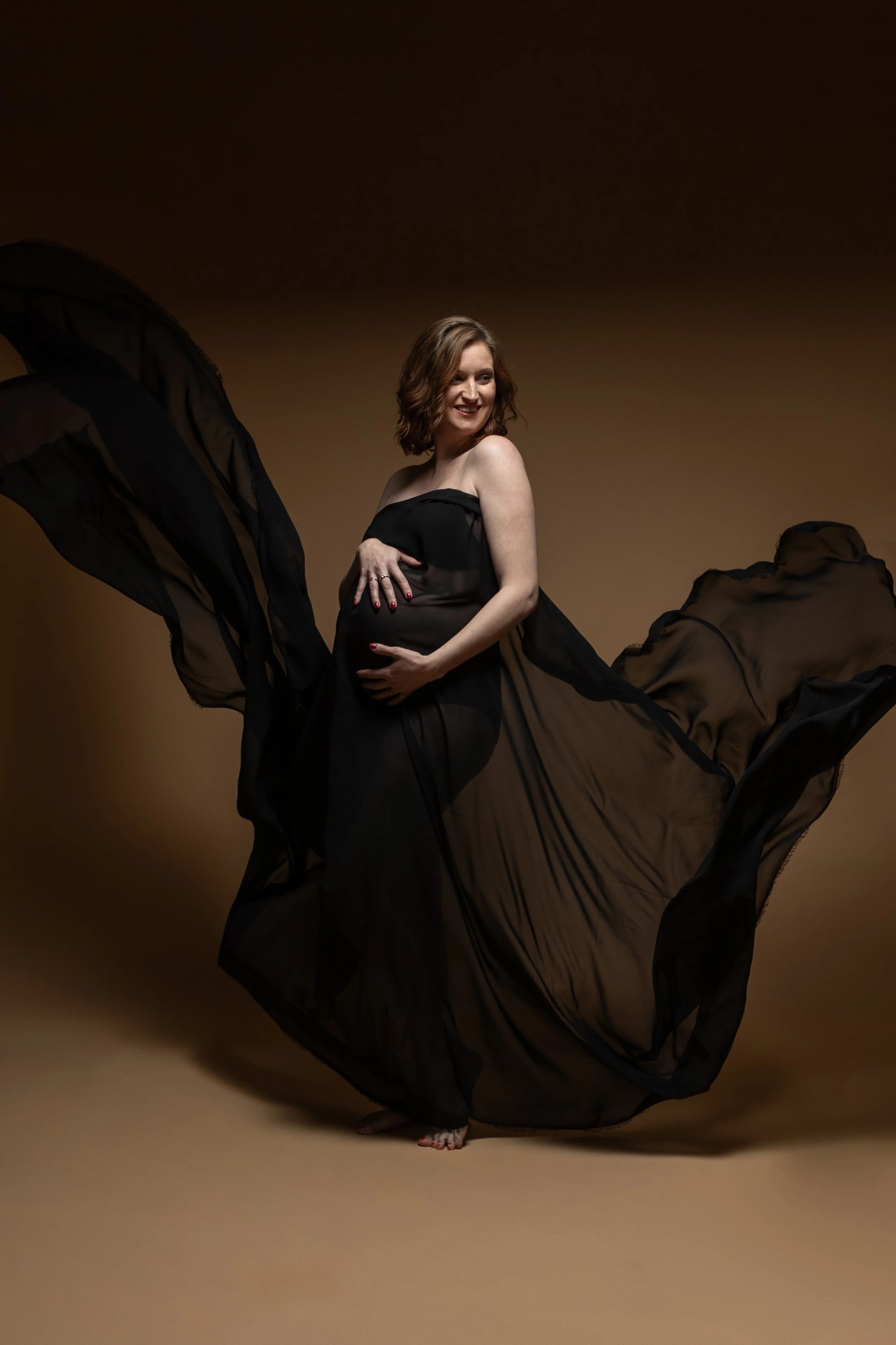 Zwangere vrouw in zwarte jurk poseert met zwart doorschijnende stof die opstijgt op een bruin achtergrond.