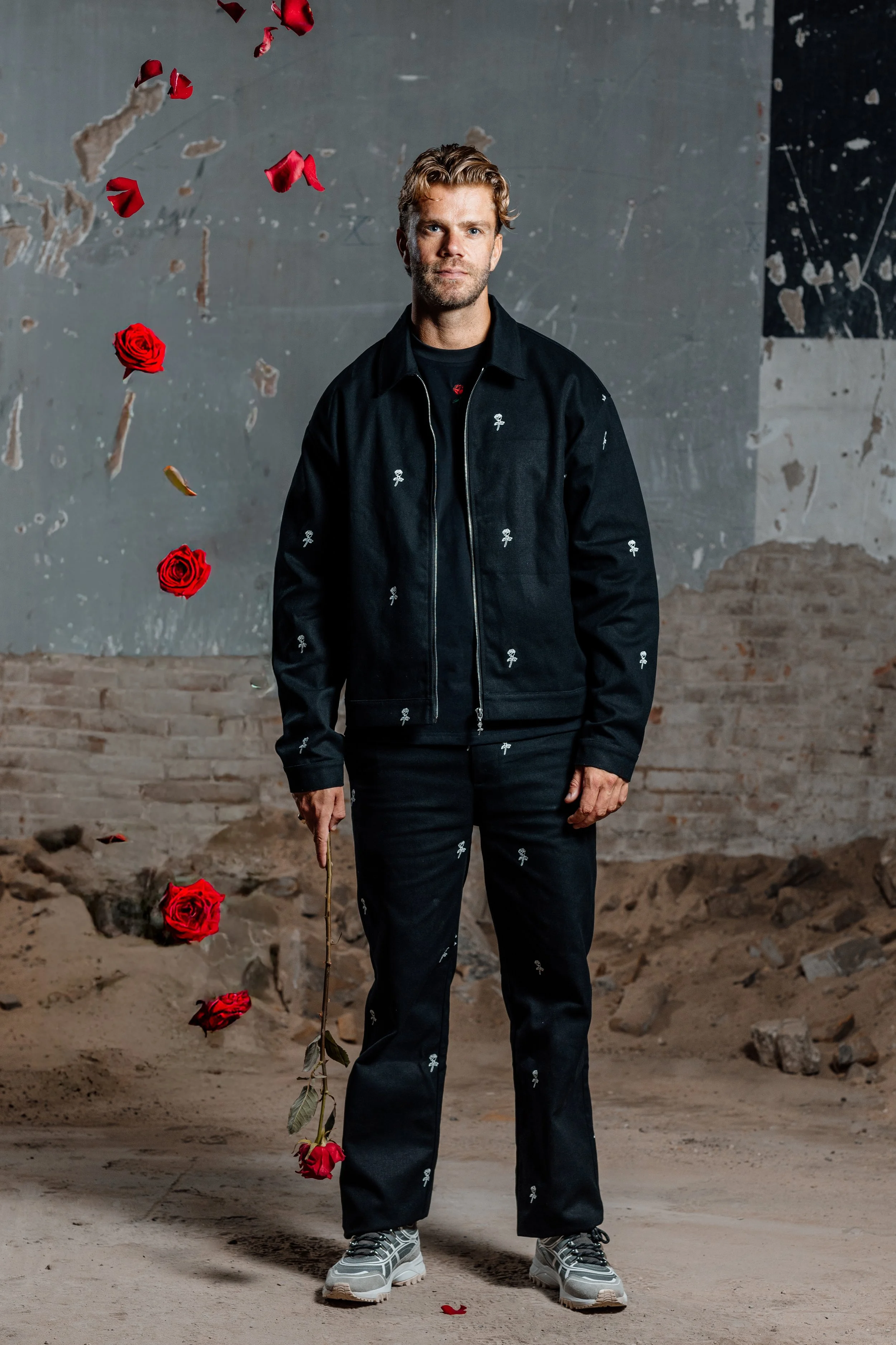 Man in zwarte outfit met patroontjes, omringd door vallende rozen en rozenblaadjes in een ruwe bakstenen ruimte.