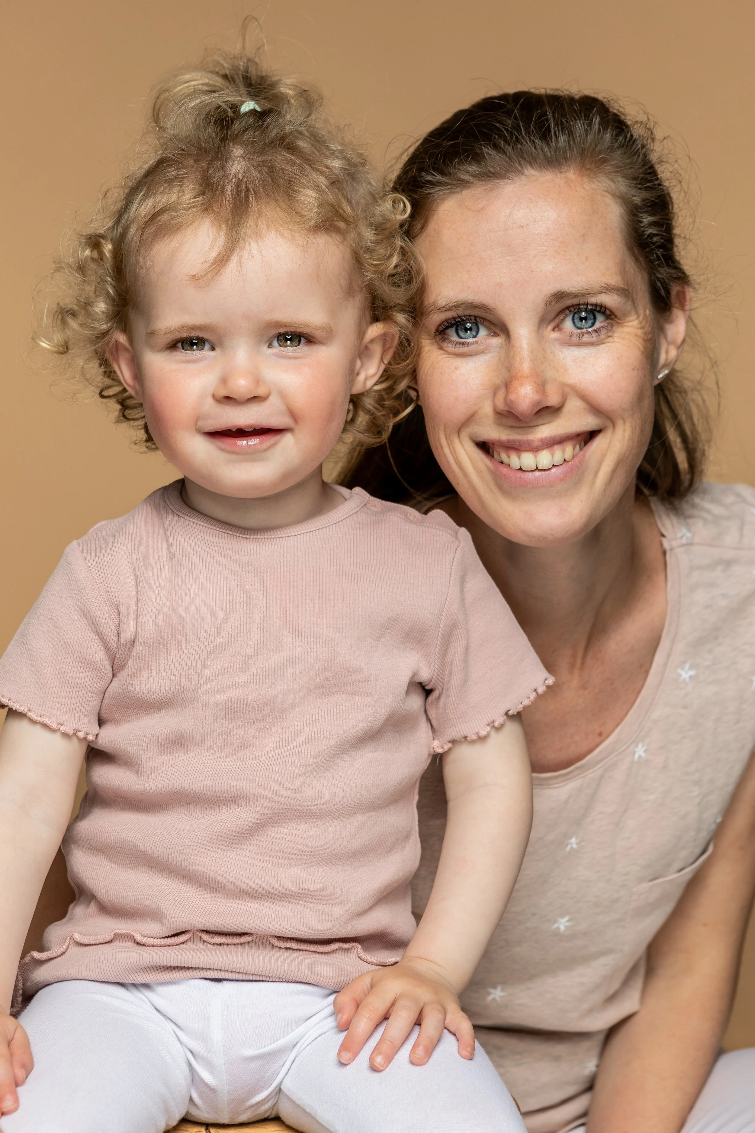 Een afbeelding van een vrolijke jonge vrouw met blond haar en blauwe ogen, zittend naast een peuter met krullend blond haar en een lichtroze shirt, beiden glimlachend 