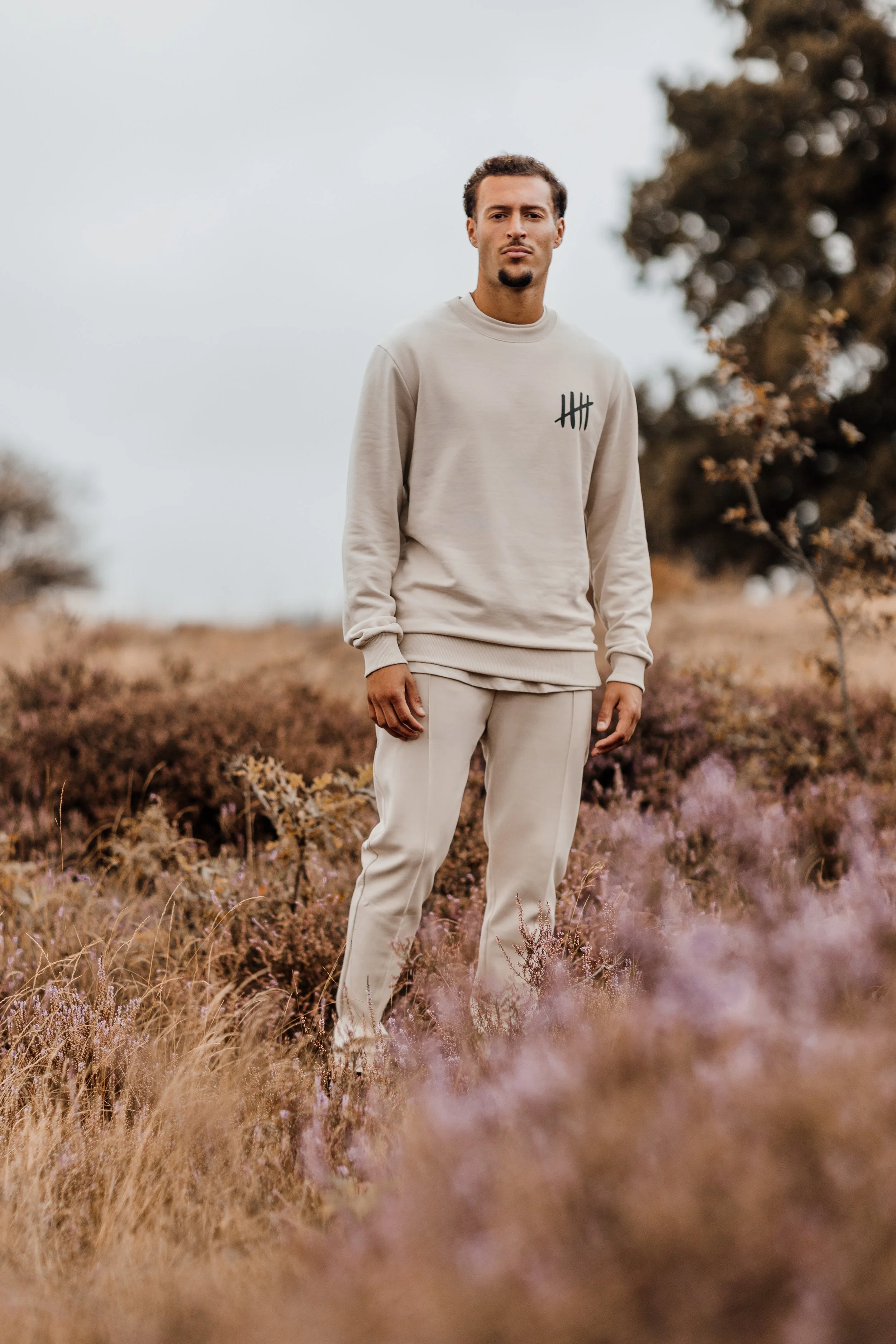 Man staat in een veld met hoge, paarse en bruine planten, draagt een beige sweatvest en joggingbroek, met een neutrale uitdrukking, onder een bewolkte hemel.