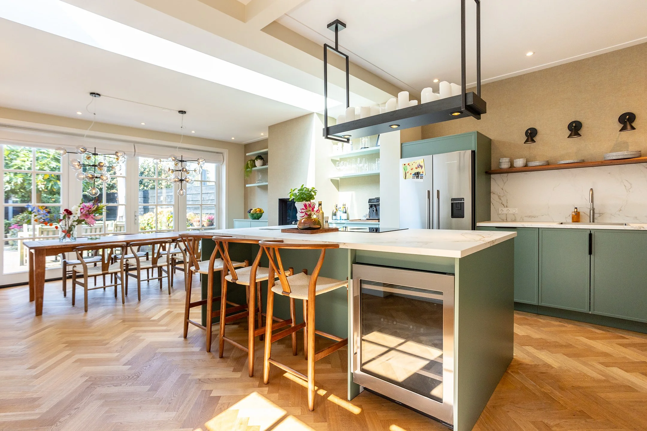 Een moderne open keuken met een eikenhouten eettafel, houten barkrukken, groene keukenkasten, een gashaard, en grote ramen die zorgen voor veel natuurlijk licht.